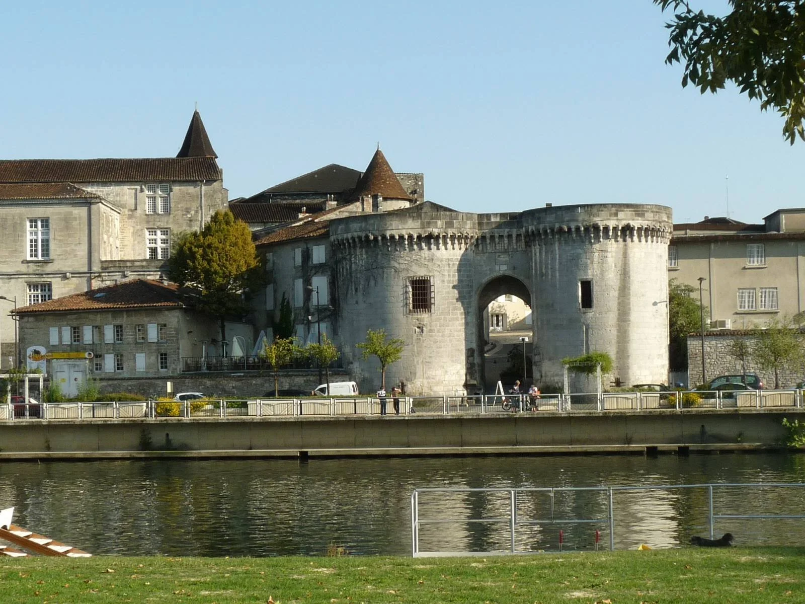 Une vieille porte fortifiée en pierre avec deux tours rondes située au bord d'une rivière, entourée de bâtiments historiques, avec un ciel clair.