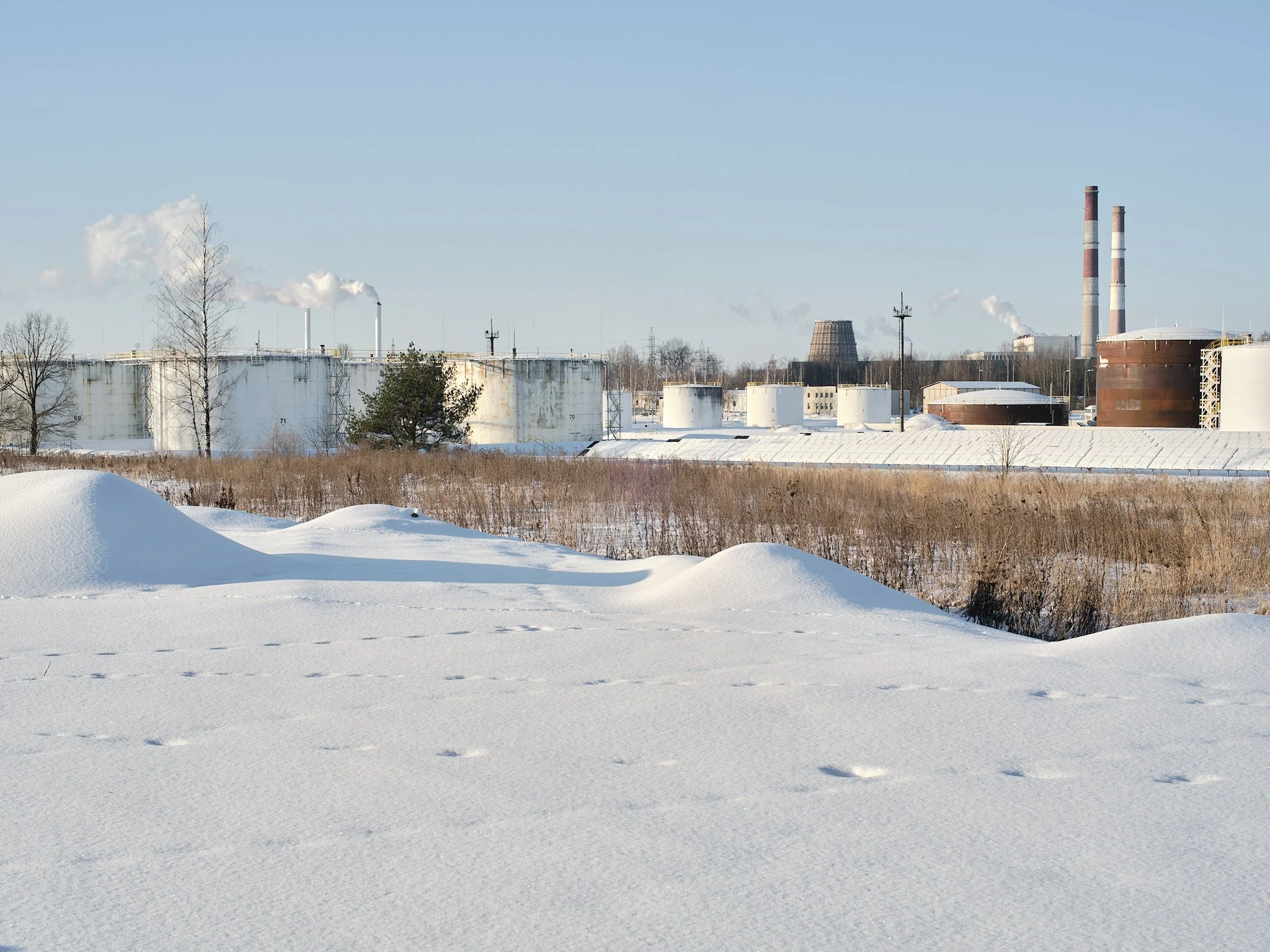 Snow-covered landscape with industrial tanks and smokestacks in the background on a clear winter day.