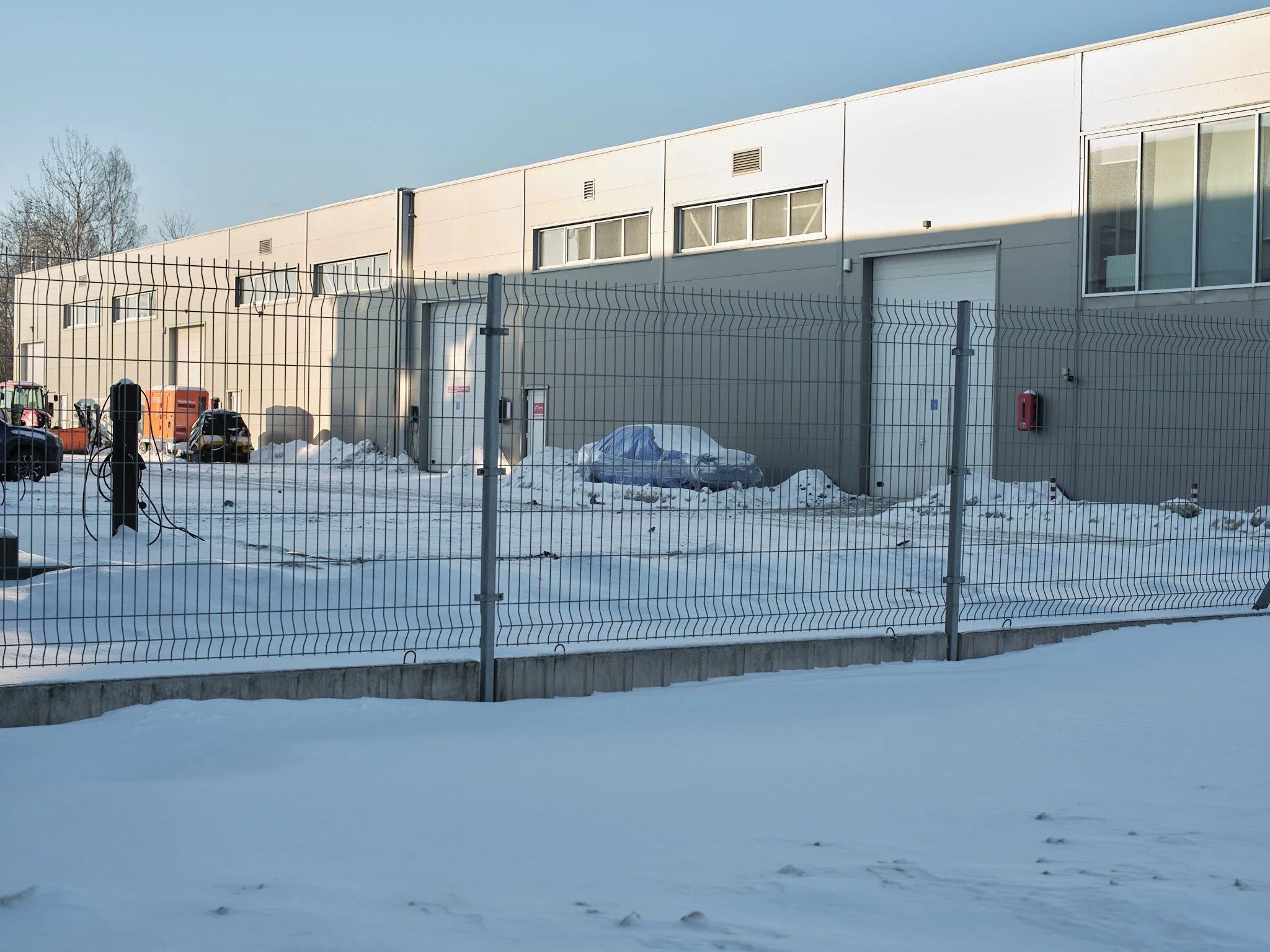 A snow-covered industrial or warehouse building with a metal fence in the foreground and parked vehicles, including a car covered with a tarp, along the side of the building.