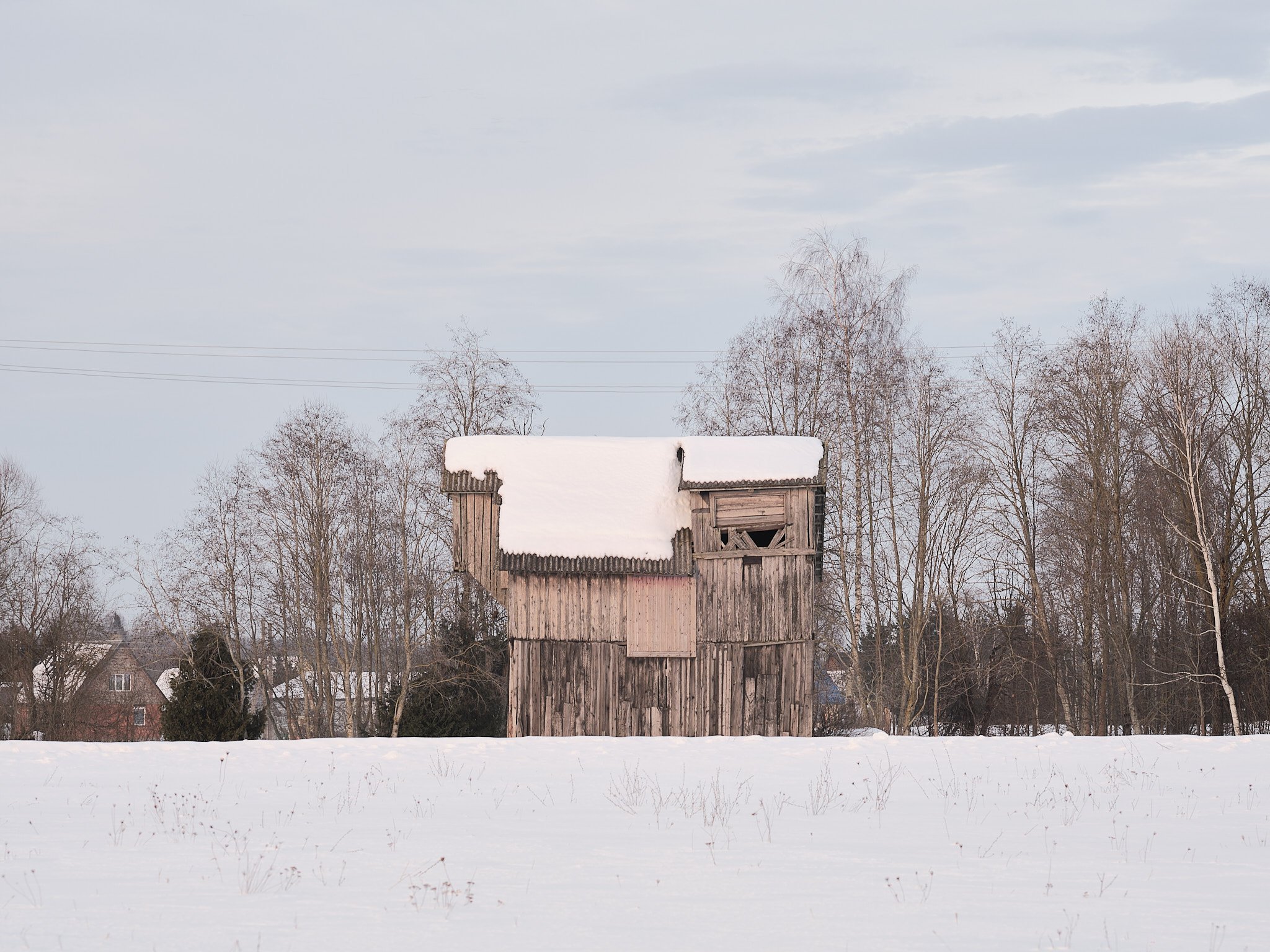 A rustic wooden barn with a snow-covered roof standing in a winter landscape with leafless trees and a snowy field.