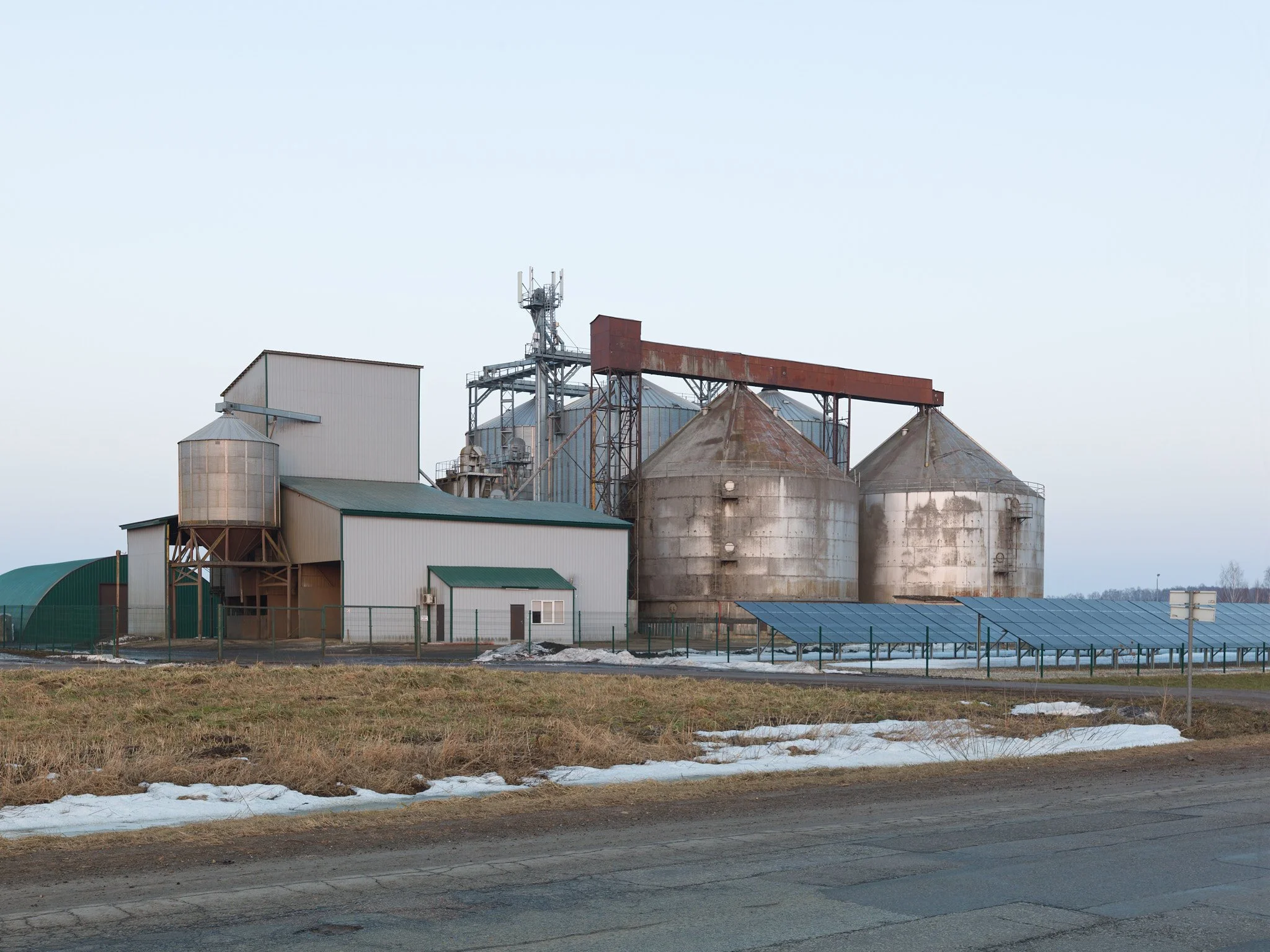 An industrial farm with grain silos and solar panels in a rural area with snow on the ground.