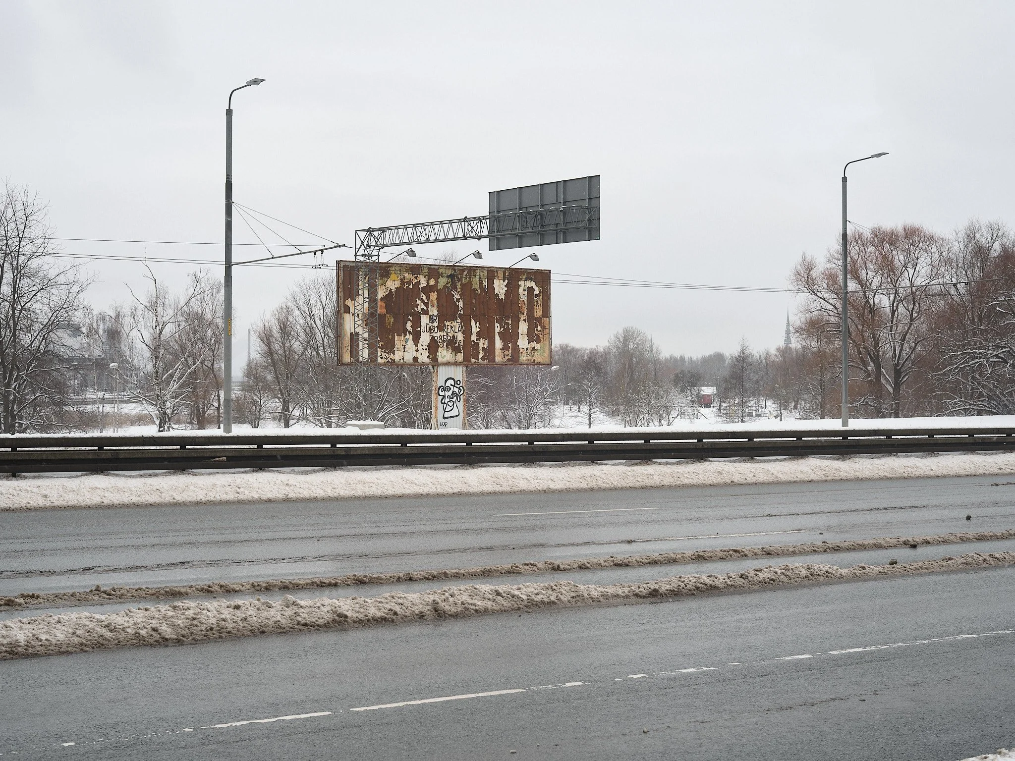 A snowy highway scene with a rusted, abandoned billboard in the distance, surrounded by leafless trees and a few houses in the background, overcast sky.