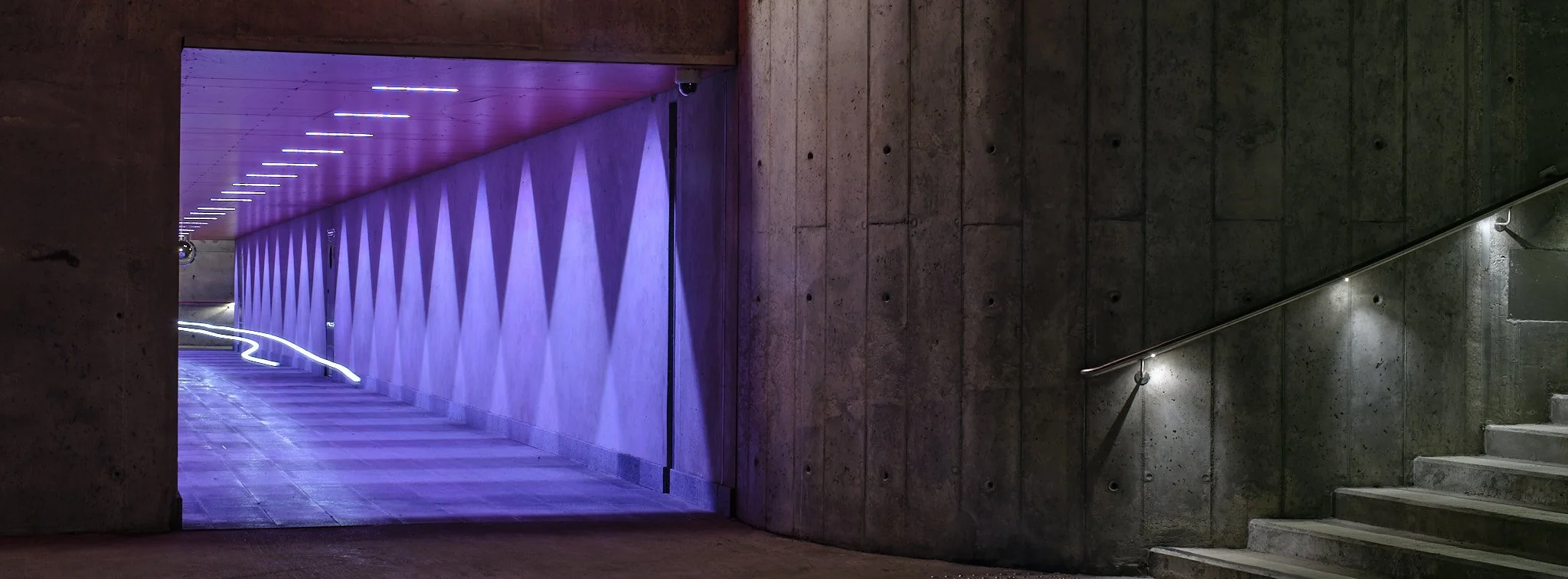 Underground pedestrian tunnel with purple and white lighting, concrete walls, and staircase on the right.