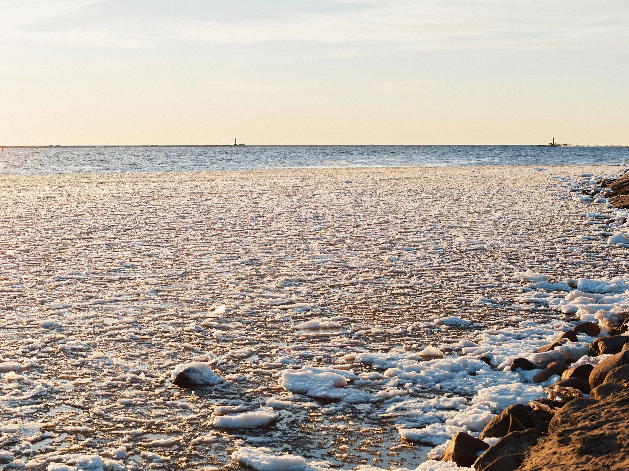 Frozen beach with ice chunks and rocks along the shoreline, with the ocean and a lighthouse in the distance during sunset.