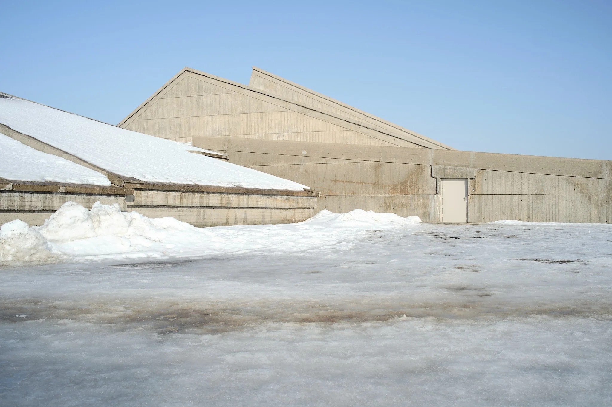 A snow-covered outdoor area with a large concrete building featuring angular rooftops, against a clear blue sky.