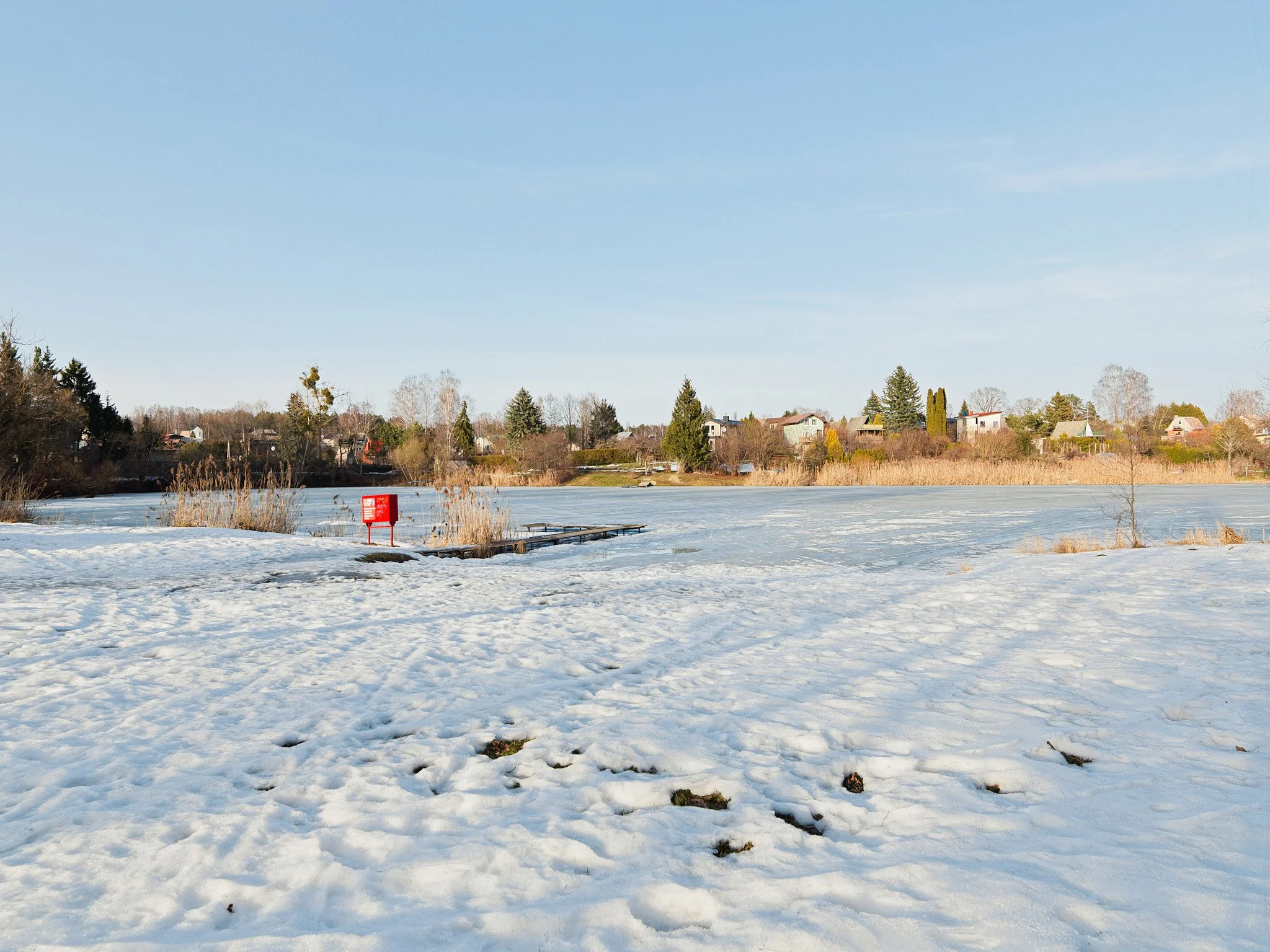Snow-covered landscape with a frozen lake, in the background are houses and trees under a clear blue sky.