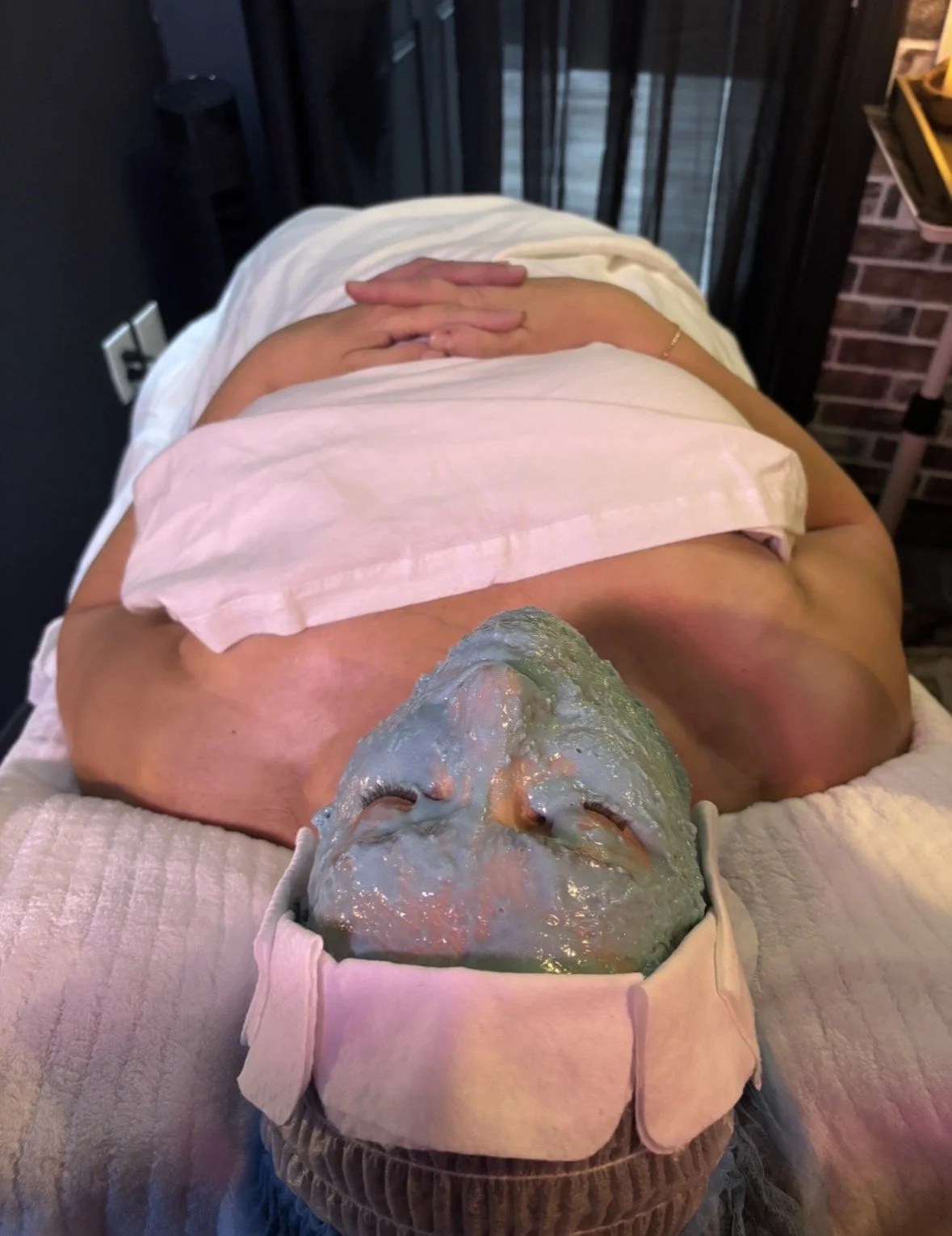 A person receiving a facial treatment at a spa or clinic, lying face up with a green mask applied to their face, while their hands are folded on their abdomen.