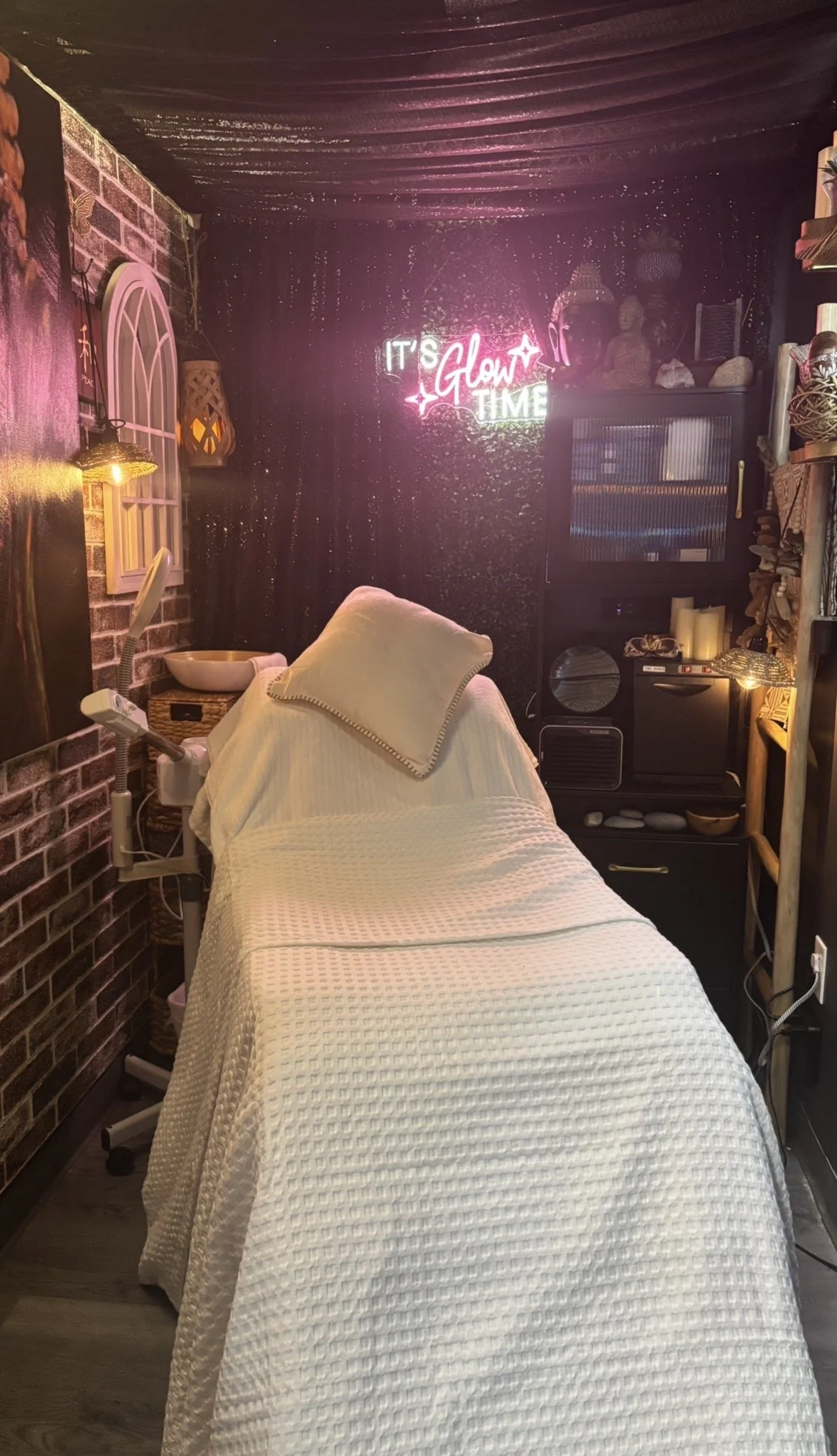 A massage or therapy room with a massage table covered in a white textured blanket and pillow. The room has dark walls, with a neon sign that reads 'It's Glow Time' in pink and white. There are decorations and shelves with candles and bottles, a small TV, and a lamp with warm lighting.