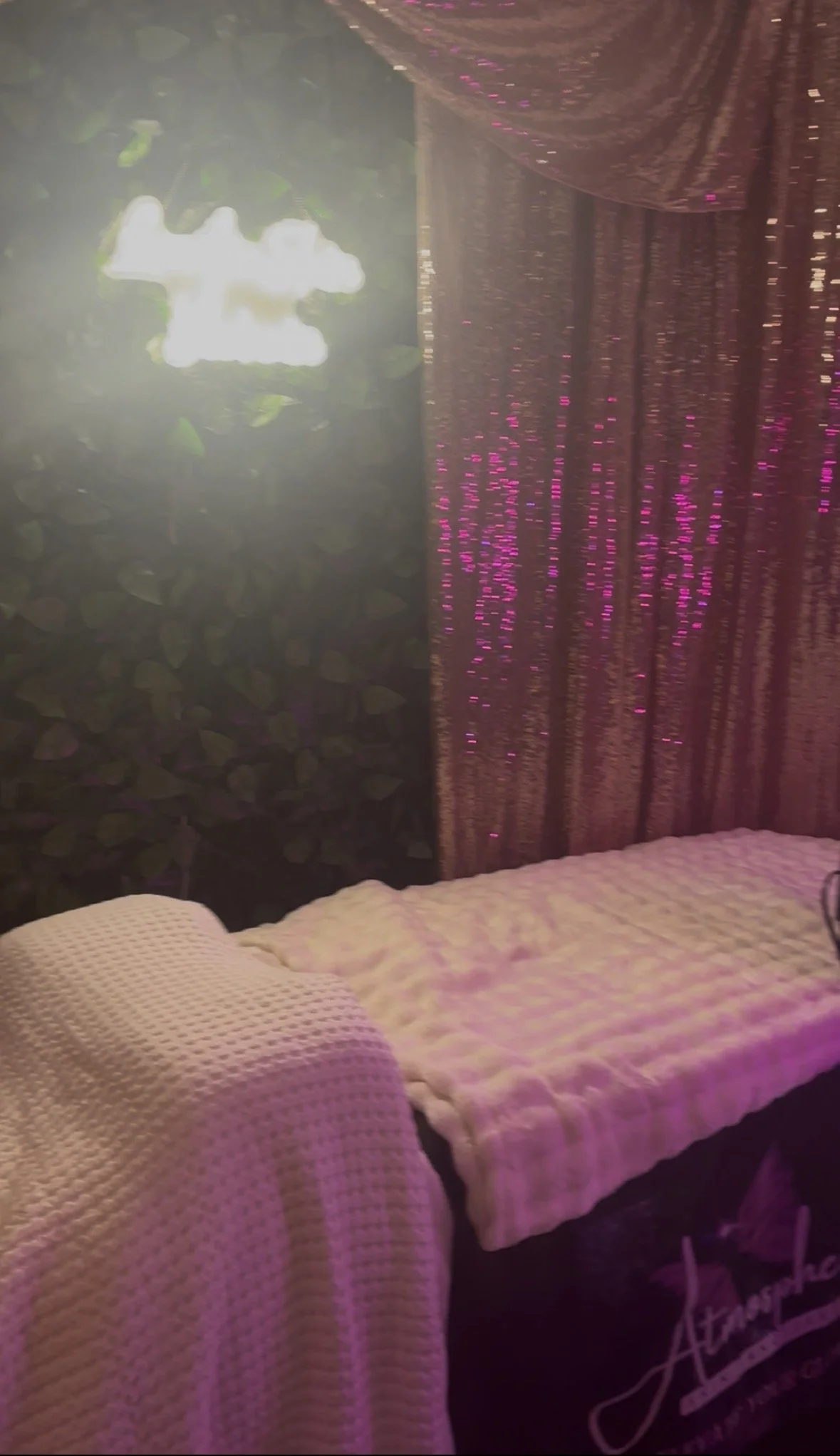 Room decorated with pink and purple lights, containing two massage tables covered with white textured blankets, one in the foreground and another in the background, with curtain backdrop and a green foliage wall.