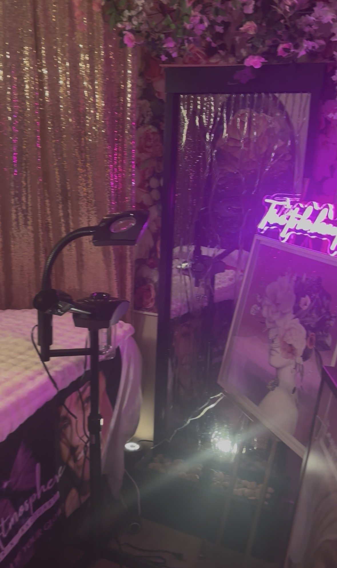 A room decorated with pink and purple flowers, a neon pink sign, a mirror, a framed portrait of a woman with flowers in her hair, and a small style lamp.