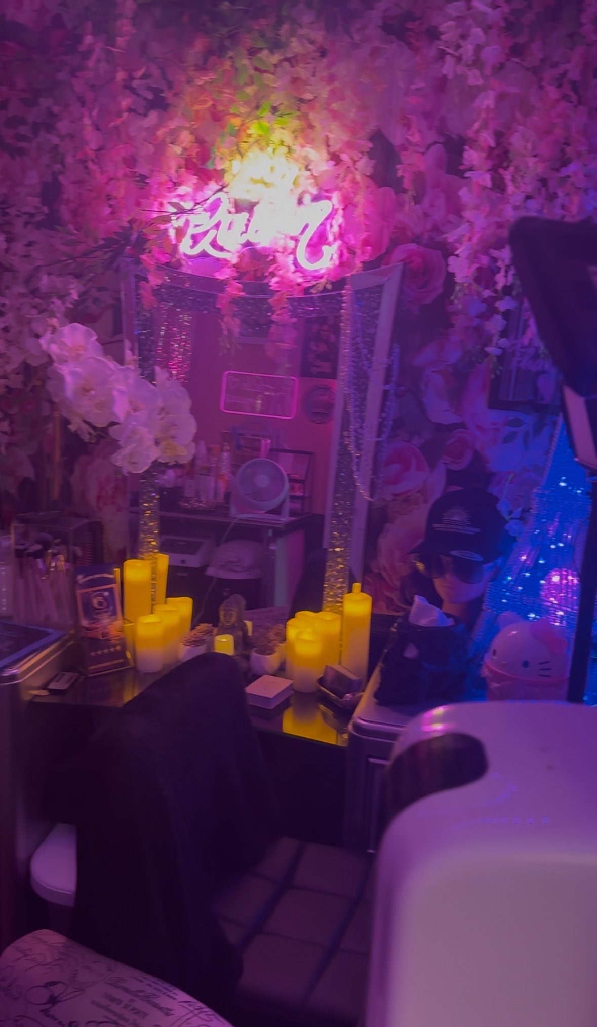 A room decorated with pink and purple flowers, illuminated by neon signs. There is a mirror, yellow candles, and various items on a desk, along with plush toys and decorative lighting creating a vibrant ambiance.