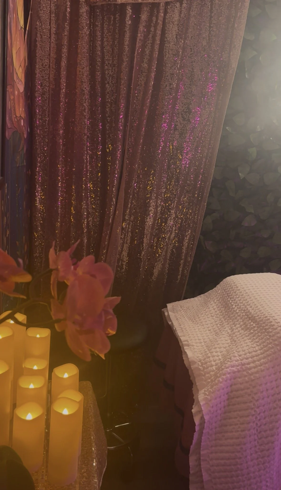 Close-up of pink flowers, several yellow LED candles, dark pink glittery curtains, and a white textured cloth covering a surface.