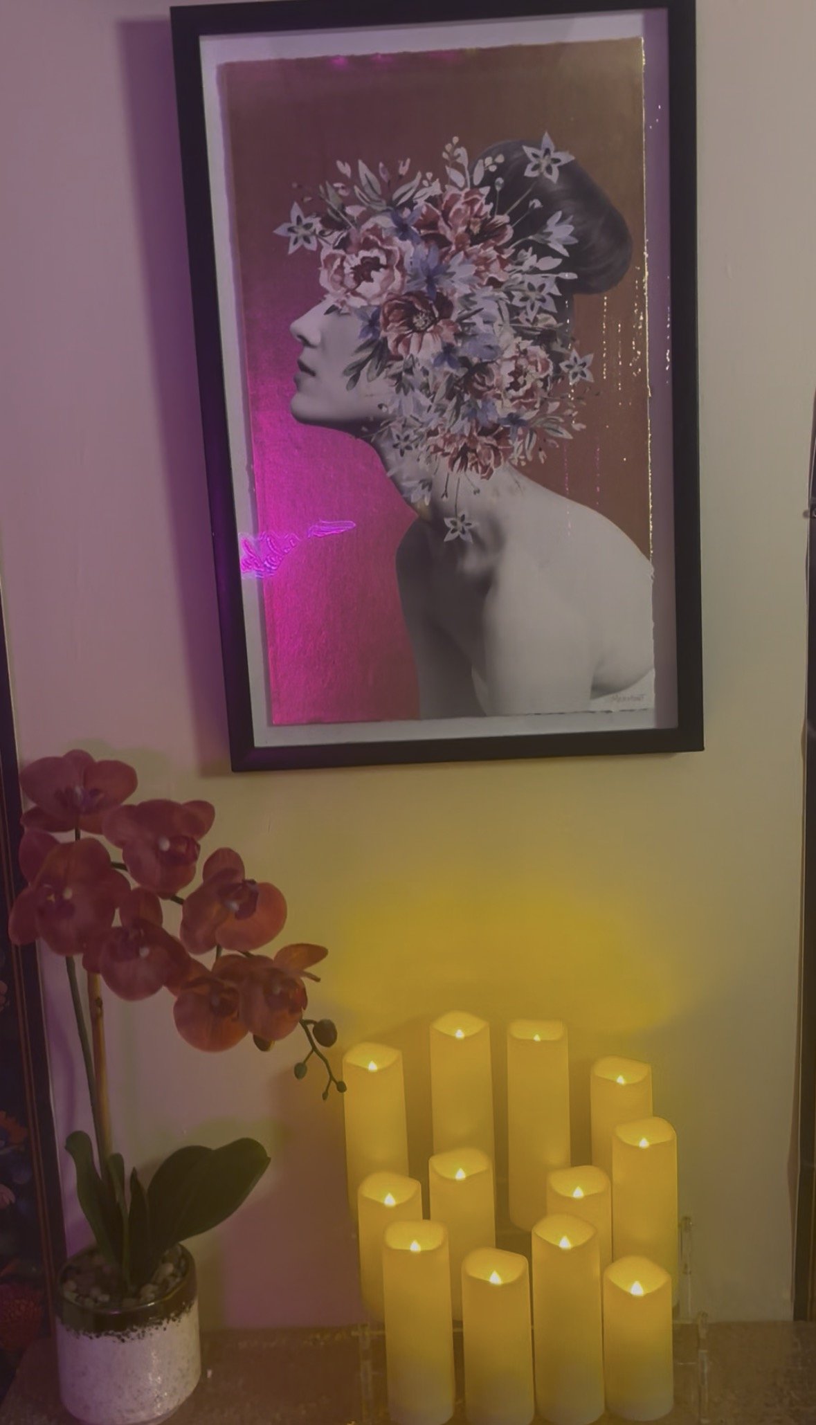 Decorative wall art featuring a black-and-white profile of a woman with a floral crown, illuminated by pink and yellow lighting, accompanied by a potted orchid and a cluster of yellow candles.