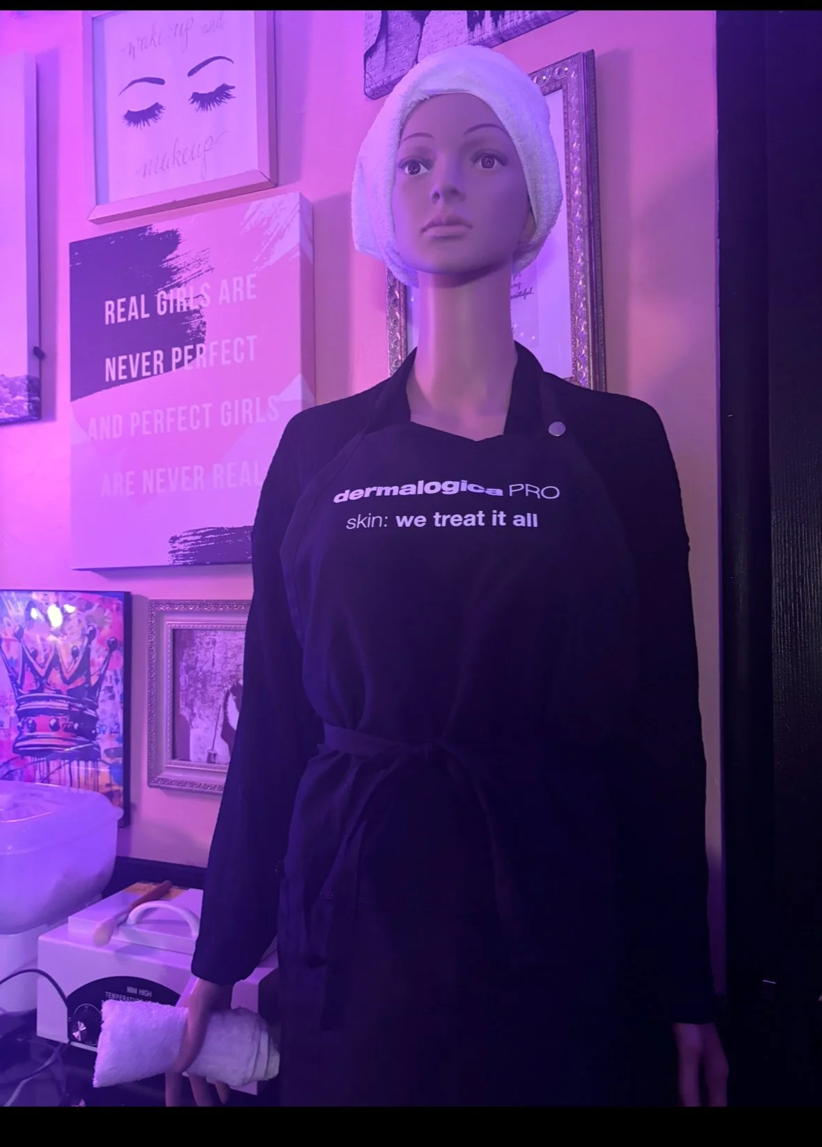 A mannequin with a towel wrapped around its head, wearing a black apron that says 'dermalogica PRO skin: we treat it all.' It is inside a room with purple lighting, with various framed artwork and signs on the wall behind it.