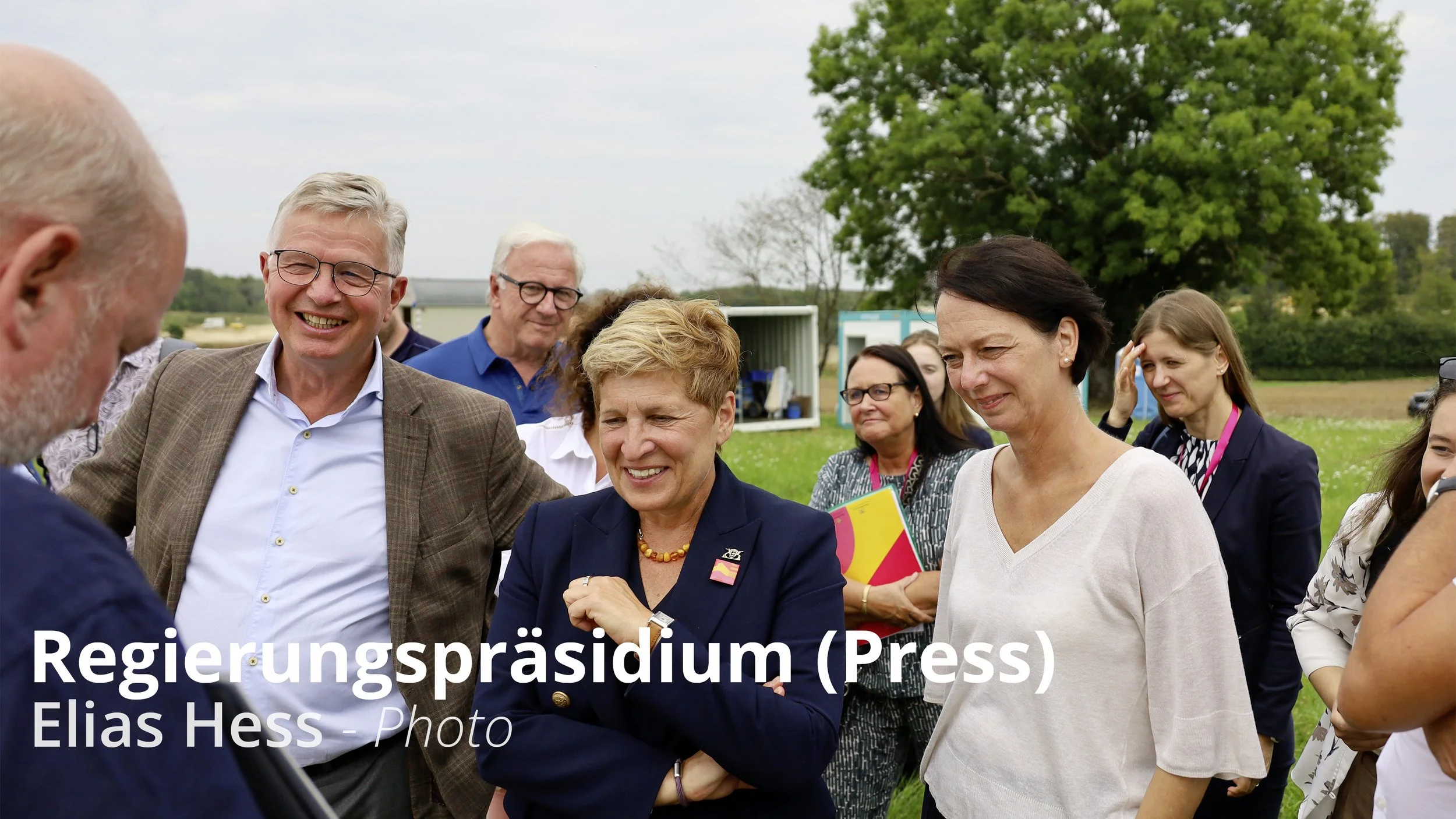 Politikerinnen und Politiker sprechen bei einem Empfang im Freien, lachen und führen Gespräche, im Hintergrund grüne Bäume und eine offene Landschaft.
