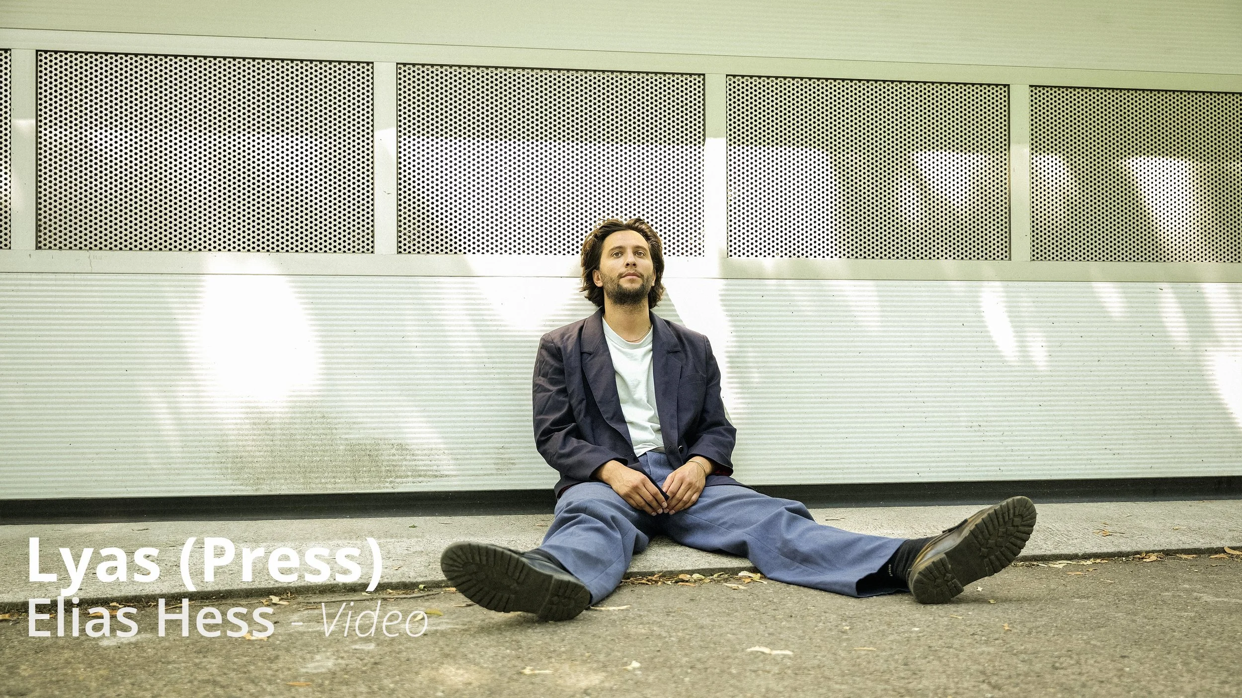 Ein Mann sitzt auf dem Boden vor einer modernen Wand mit perforierten Metallplatten. Er trägt eine dunkle Jacke, eine weiße T-Shirt und große, lockere Hosen. Der Mann hat dunkle, medium-lange Haare und einen Bart.