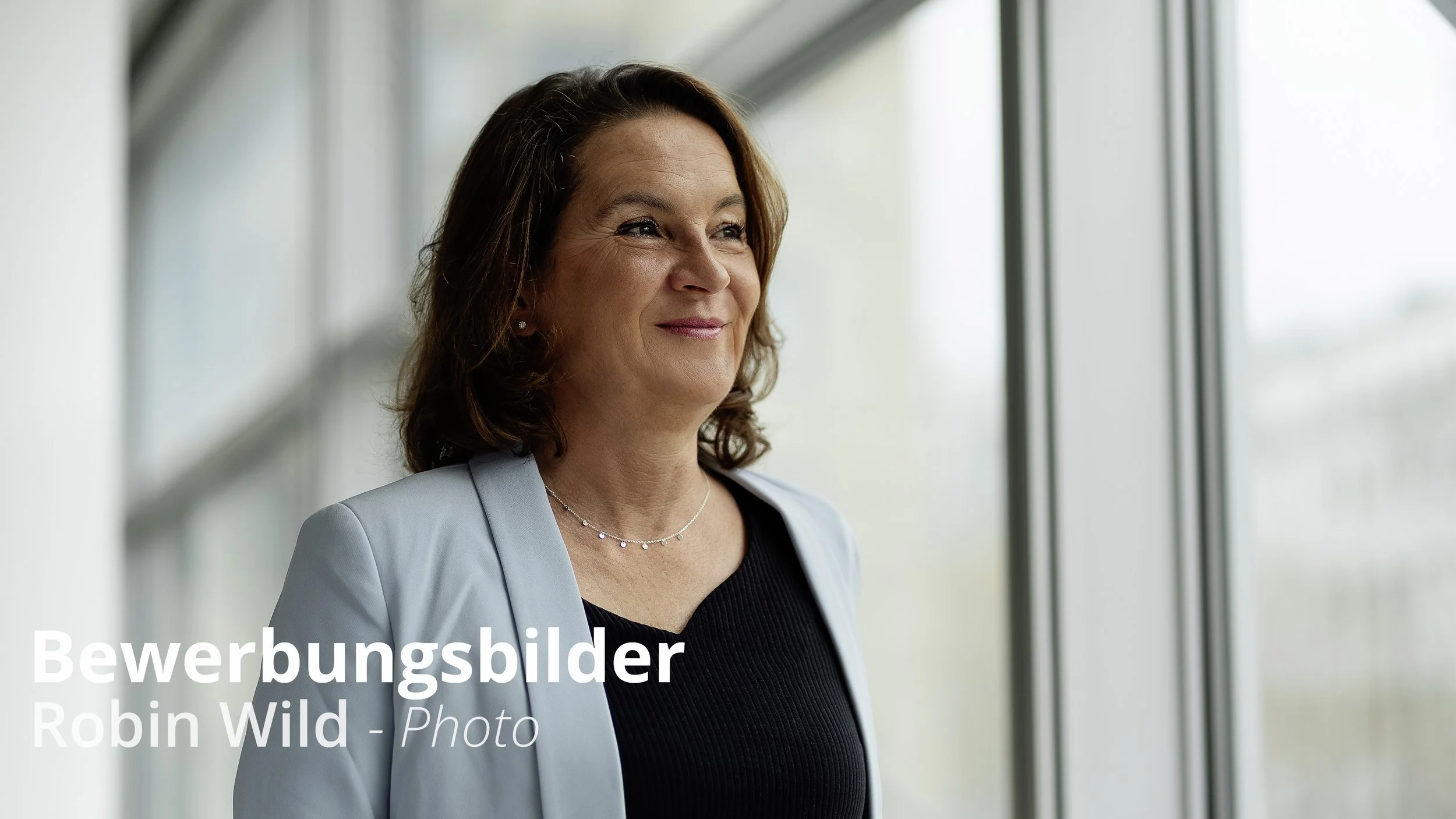 Eine lächelnde Frau mit braunen, schulterlangen Haaren steht vor einem großen Fenster in einem modernen Gebäude. Sie trägt eine hellblaue Jacke, ein schwarzes Oberteil und eine dünne Halskette.