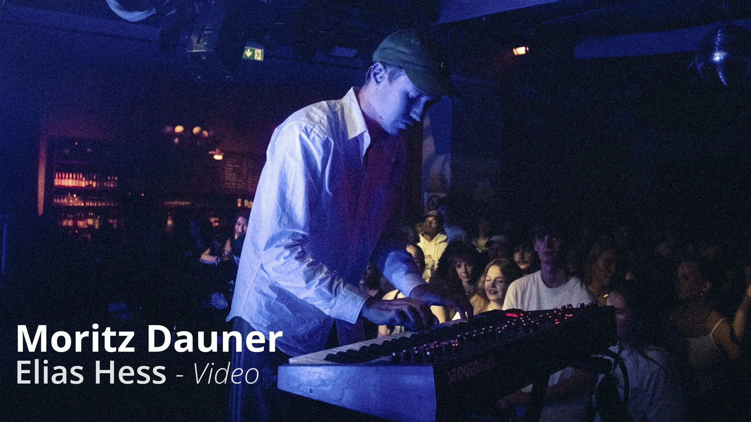 Ein Musiker spielt auf einem Keyboard in einem dunklen Club, umgeben von einem Publikum, das zuschaut.