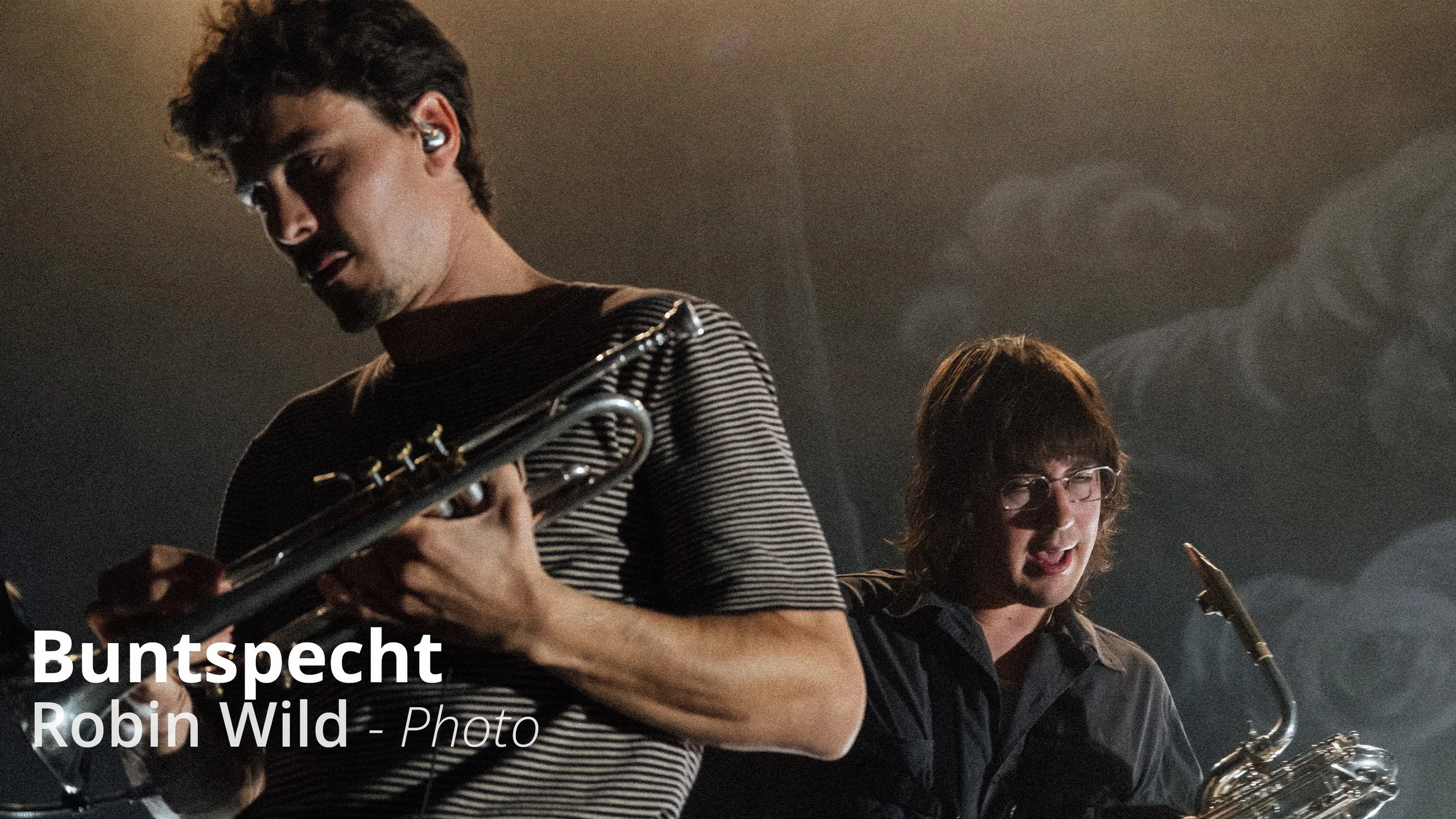 Zwei Musiker (Die Band Buntspecht) spielen Saxophon und Trompete bei einem Live-Konzert, einer trägt ein gestreiftes Hemd, der andere eine dunkle Jacke, im Hintergrund dunkle Bühne mit Rauch.