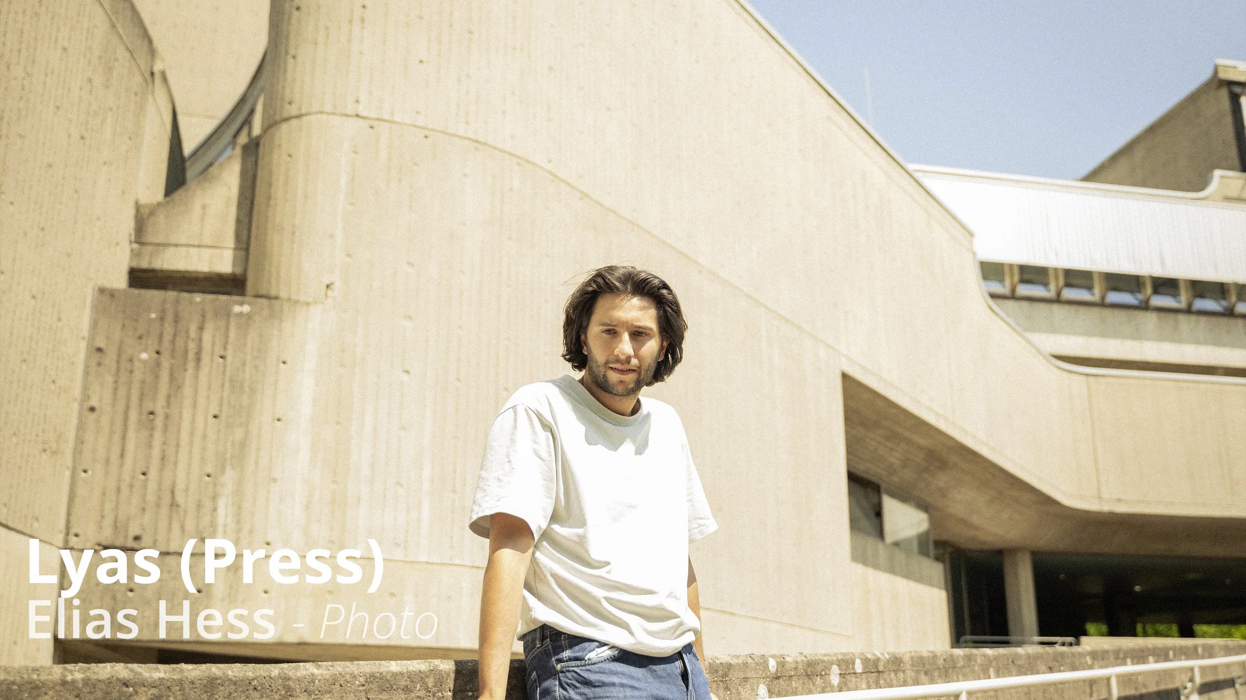 Ein junger Mann mit braunen Haaren trägt ein weißes T-Shirt und blaue Jeans und steht vor einem modernen, beige Betonstrukturgebäude bei sonnigem Wetter.