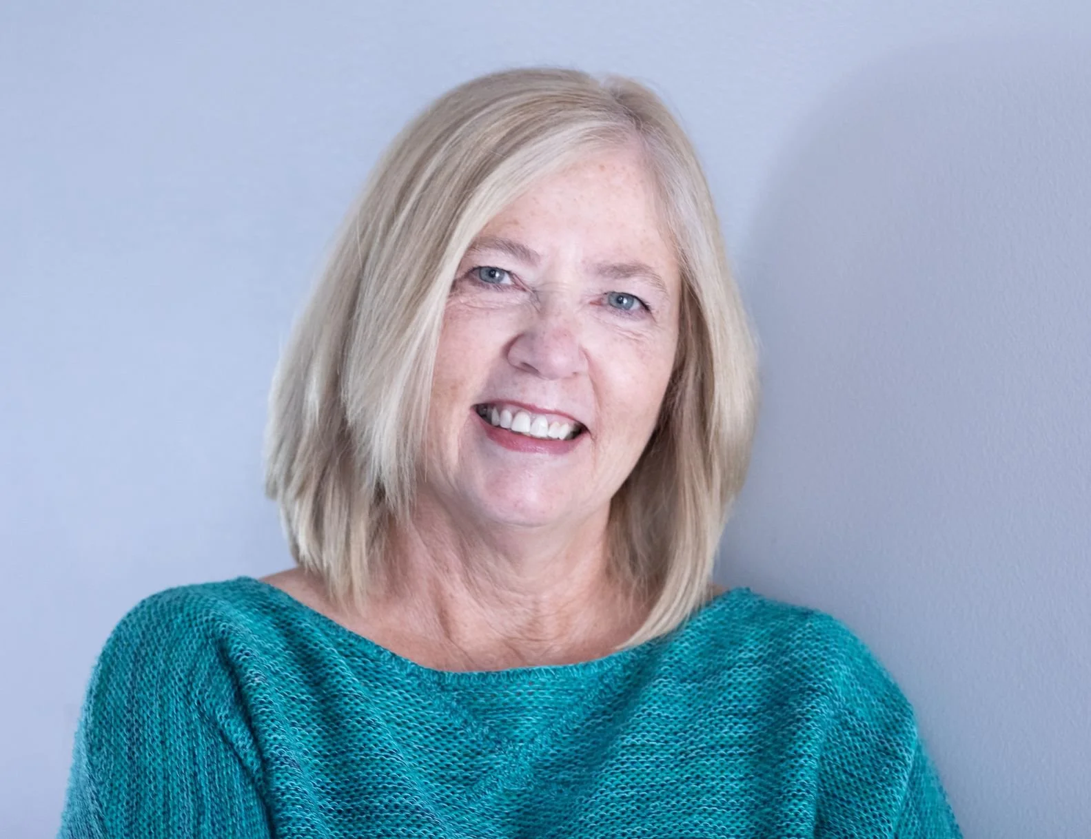 Portrait of a smiling middle-aged woman with blonde hair wearing a teal sweater in front of a plain gray background.