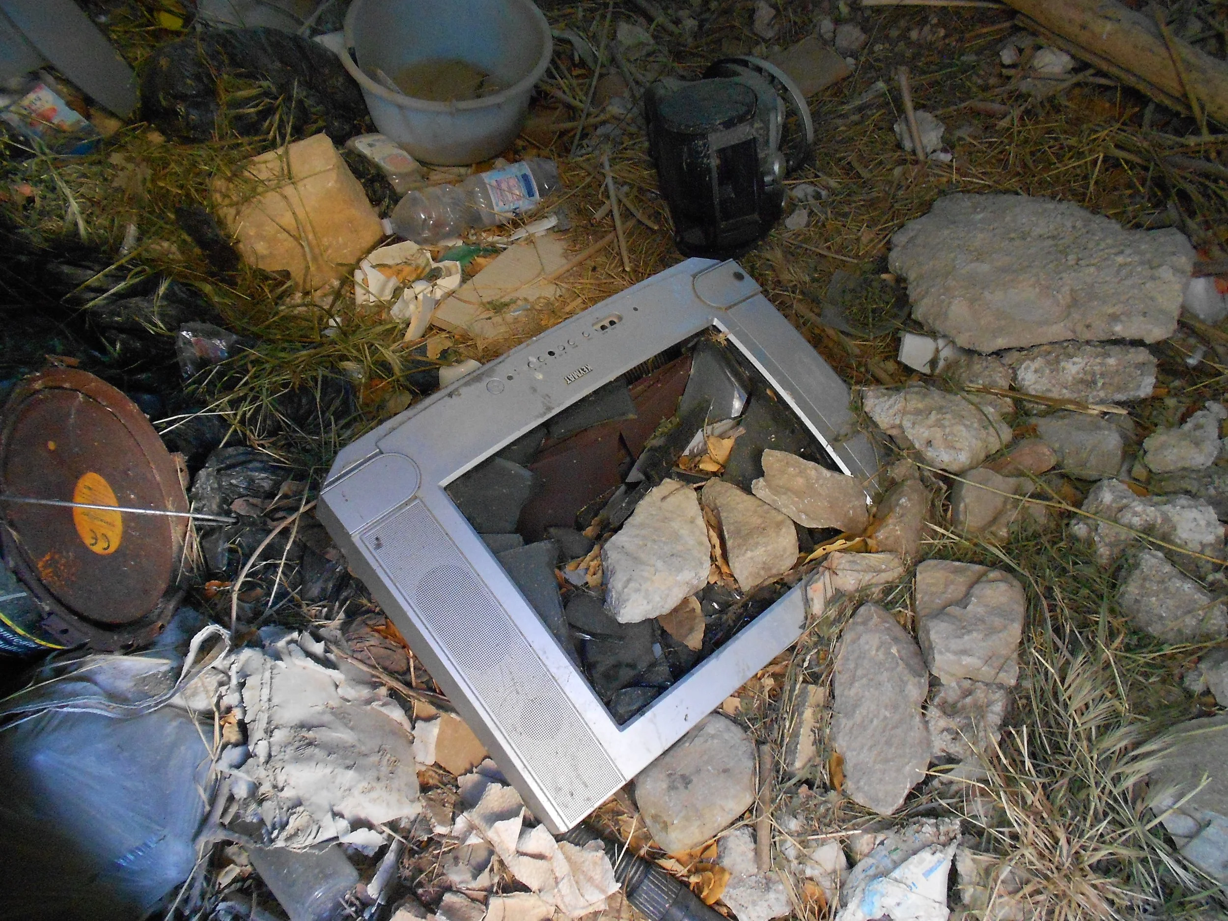 Débris et déchets électroniques, dont un écran cassé, entourés de pierres, plastique, boîtes en carton, et autres détritus