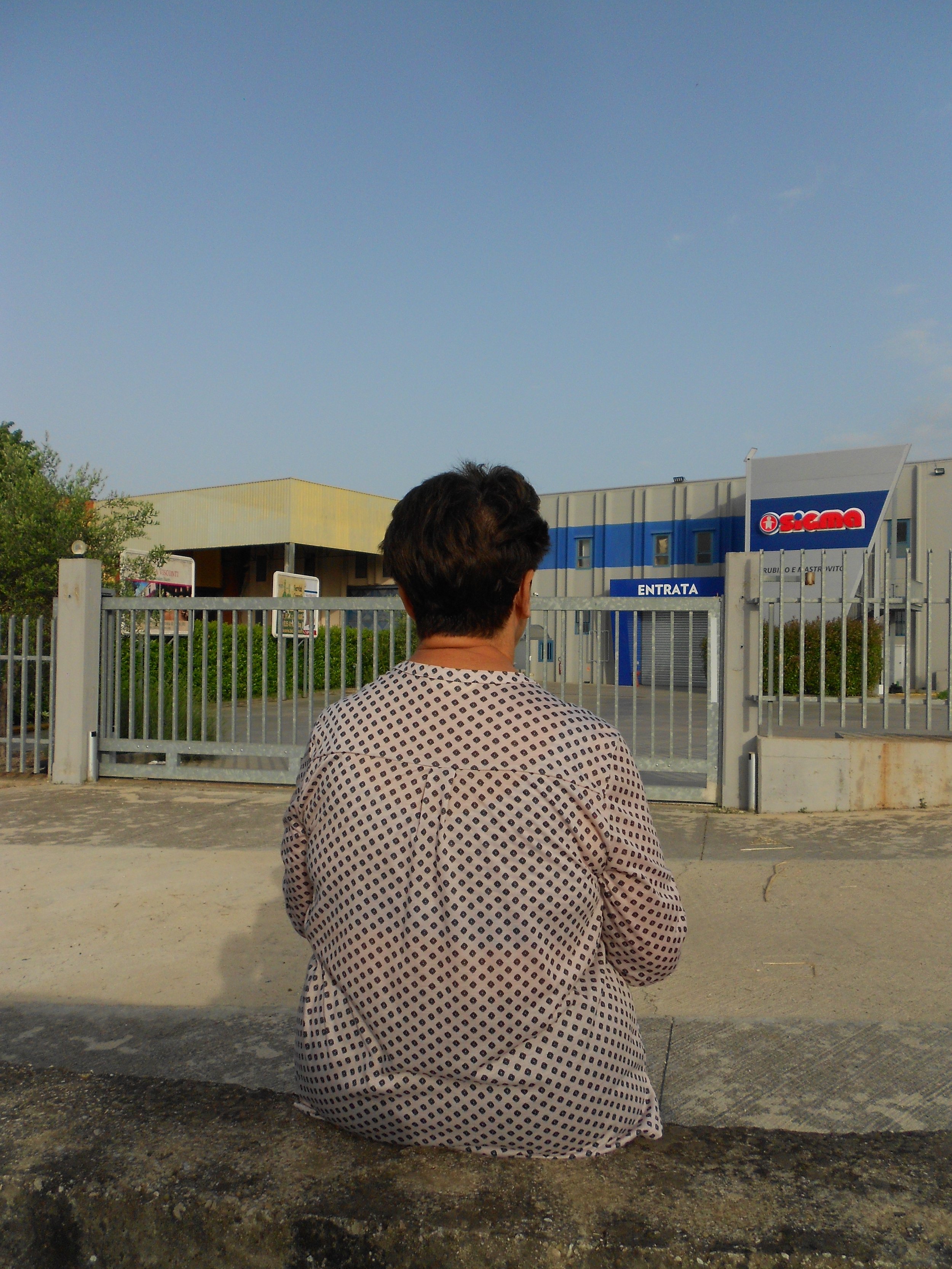 Une personne assise devant une clôture métallique, regardant une entrée en usine ou center commercial, avec un bâtiment en arrière-plan et un ciel clair.