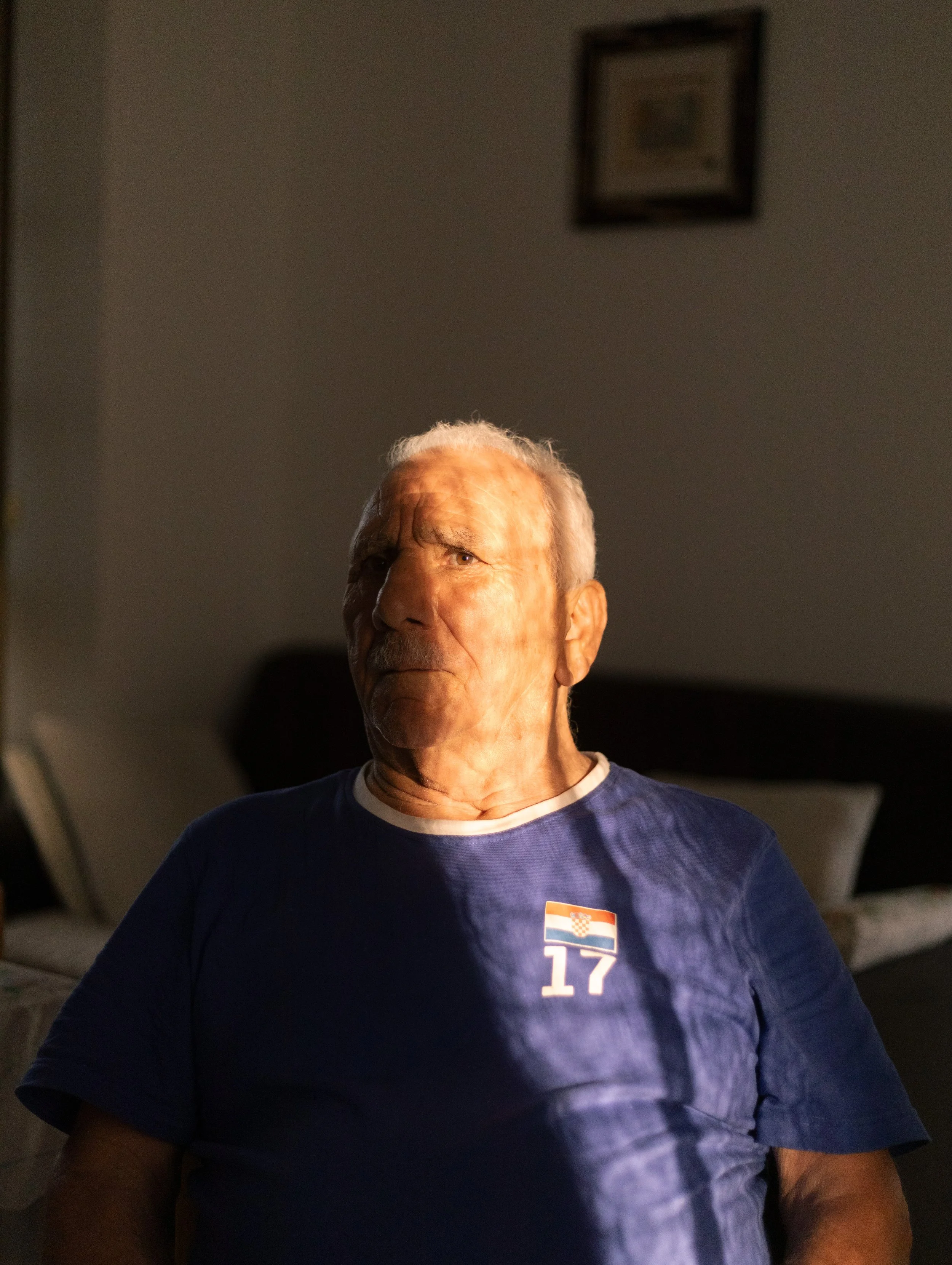Un homme âgé assis dans une pièce sombre, éclairé par la lumière naturelle, portant un t-shirt bleu avec le numéro 17 et un drapeau croate.