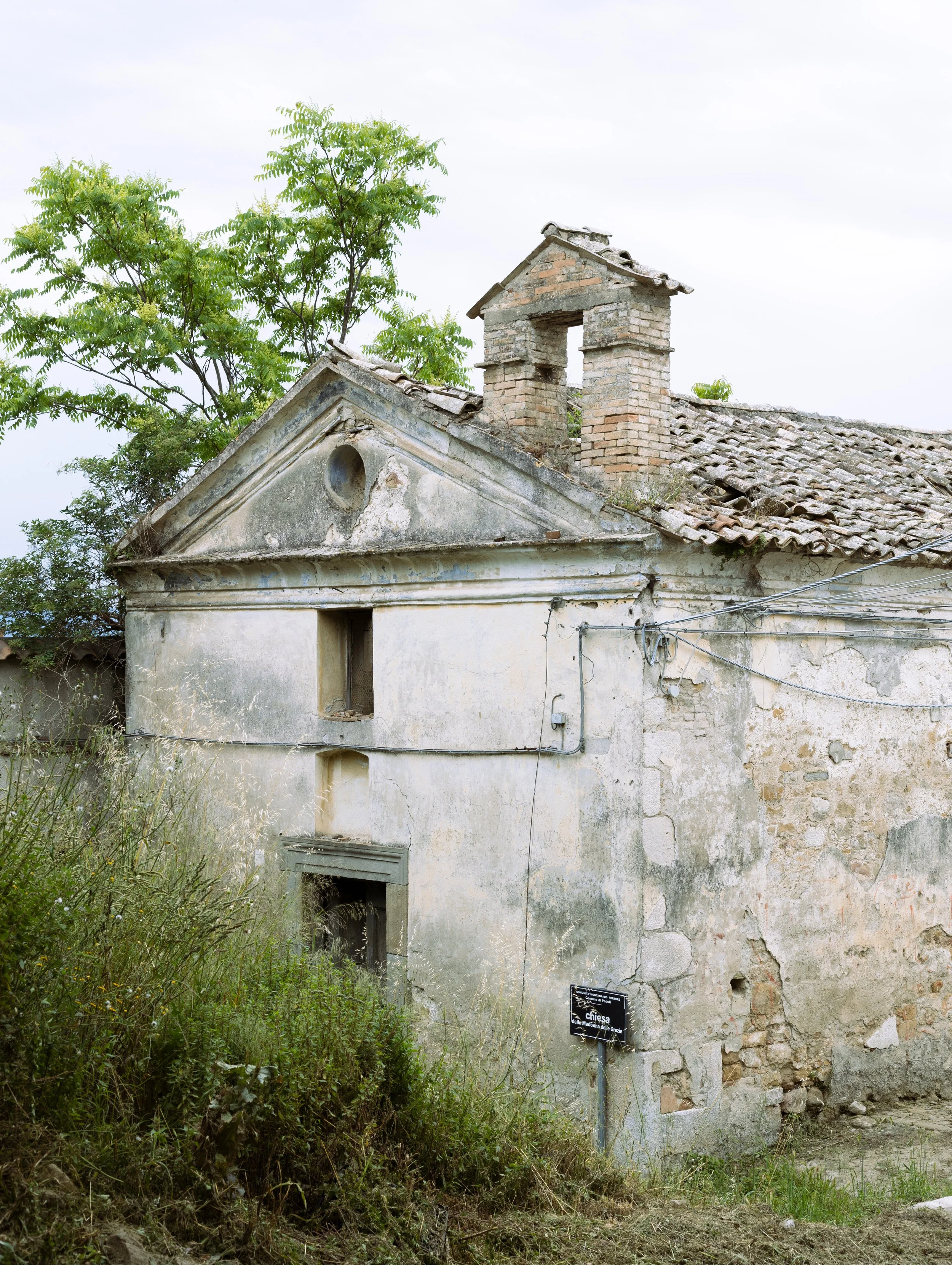 Une vieille église en pierre avec un toit en tuiles, entourée de végétation, et un panneau indiquant 'Chiesa'.