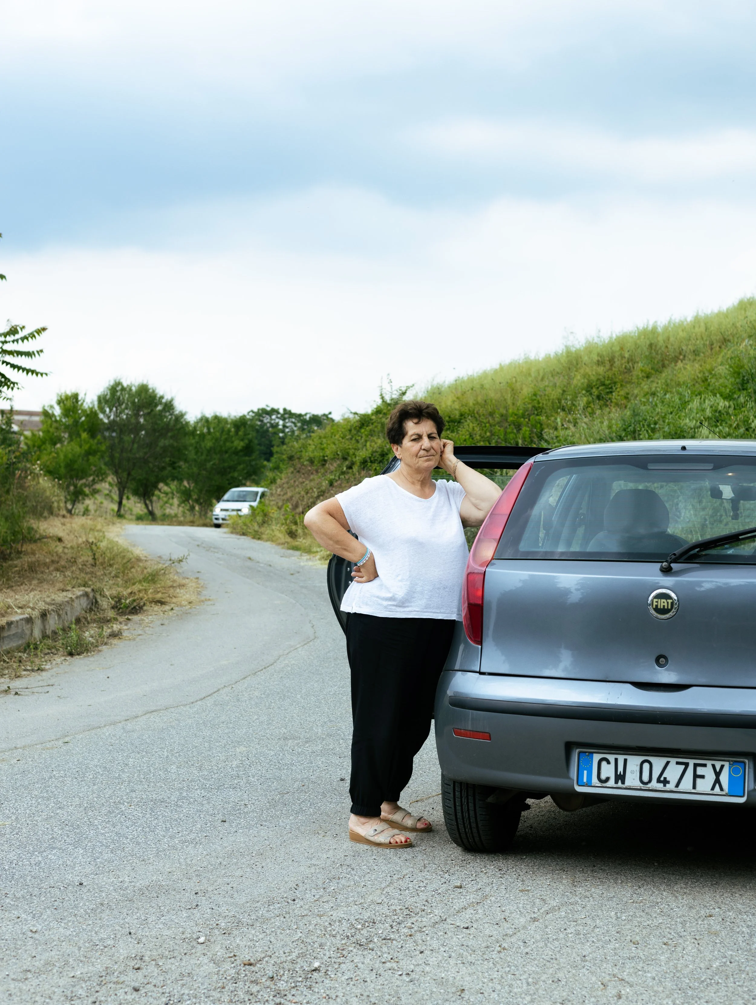 Femme âgée à côté d'une voiture grise sur une route rurale, avec un paysage verdoyant et un ciel nuageux en arrière-plan.