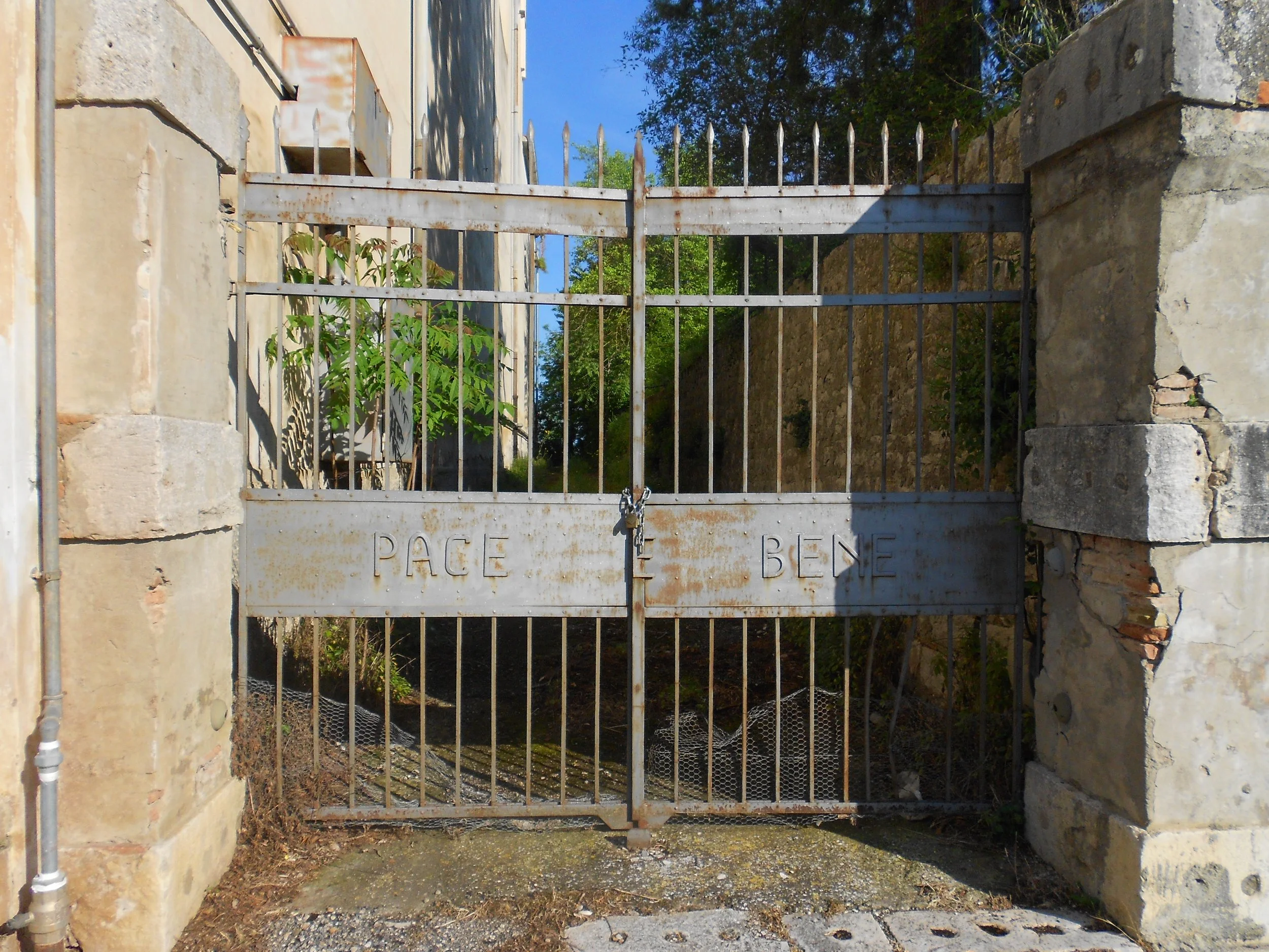 Une grille en métal rouillée verrouillée entre deux piliers en pierre, avec l'inscription 'PLACE' à gauche et 'BENE' à droite, entourant un espace avec des plantes et un arbre en arrière-plan.