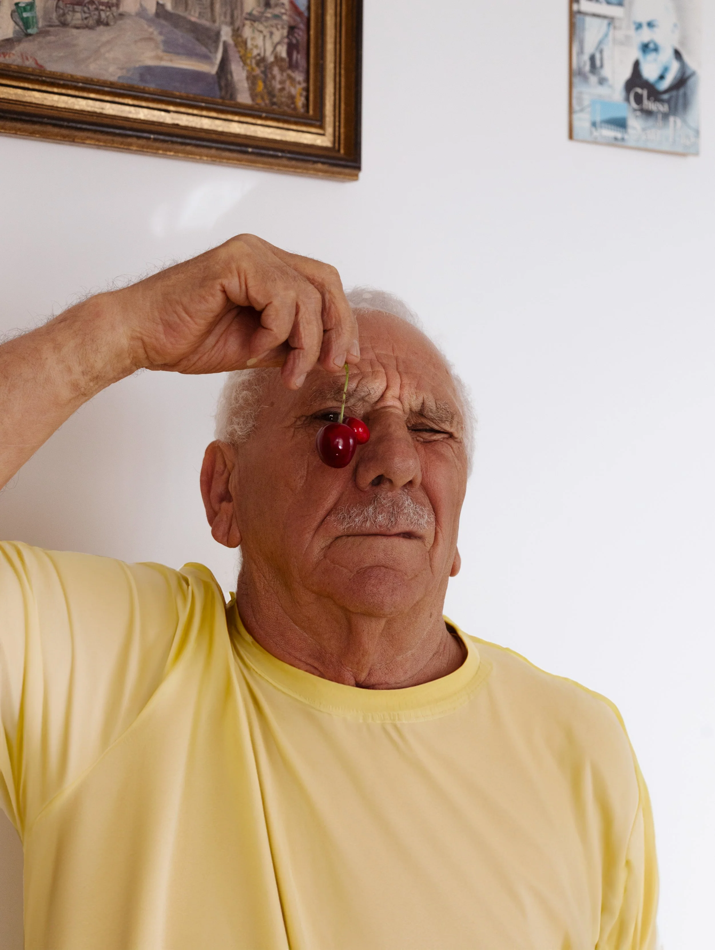 Un homme âgé, portant un t-shirt jaune, tient une cerise devant un œil fermé, posée sur son front, dans une pièce avec des tableaux au mur.
