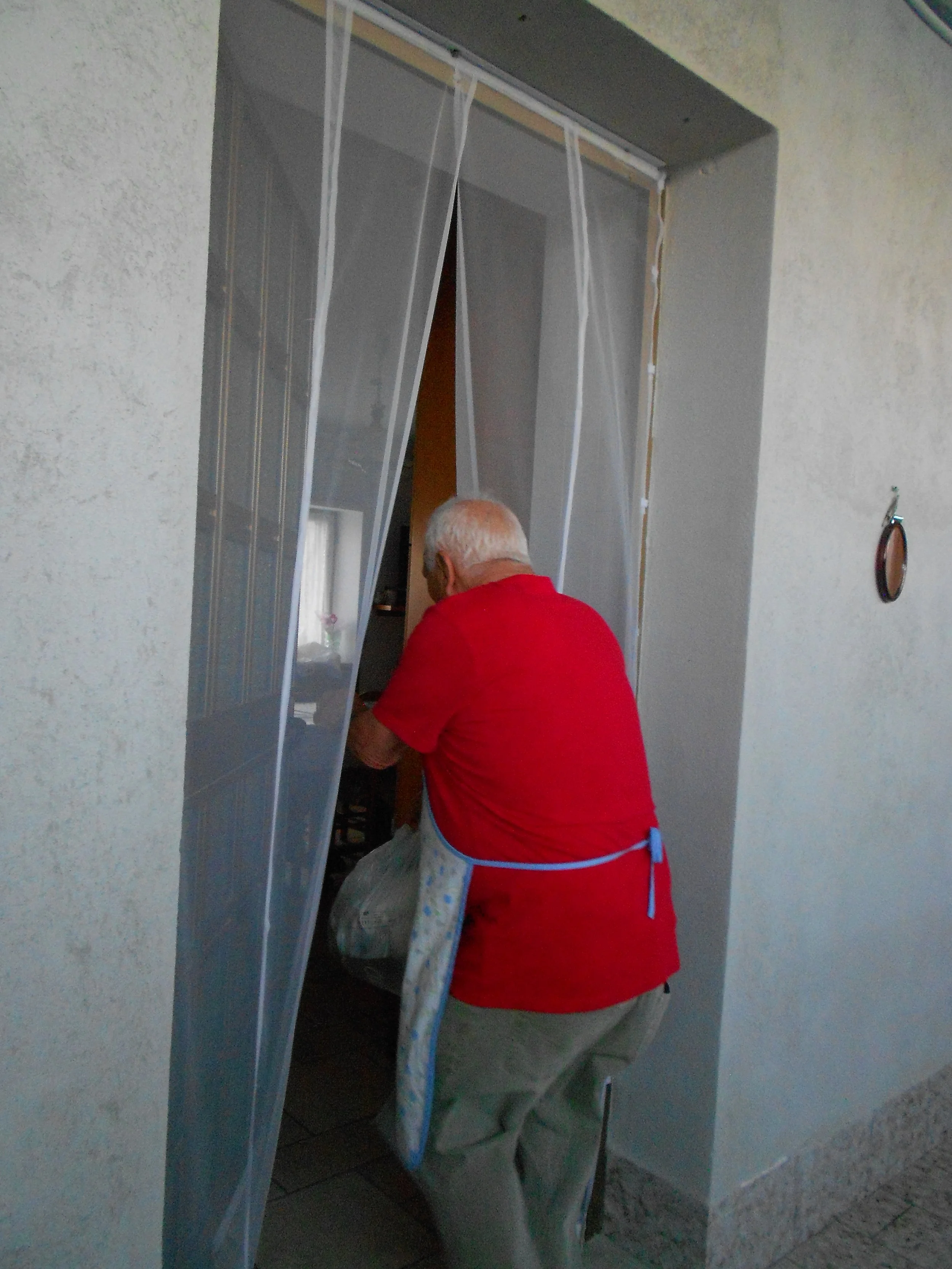 Un homme âgé, portant un t-shirt rouge et un tablier, se tient dans l'entrée d'une maison en tenant un sac. L'entrée est partiellement barricadée avec des bâches en plastique transparent.