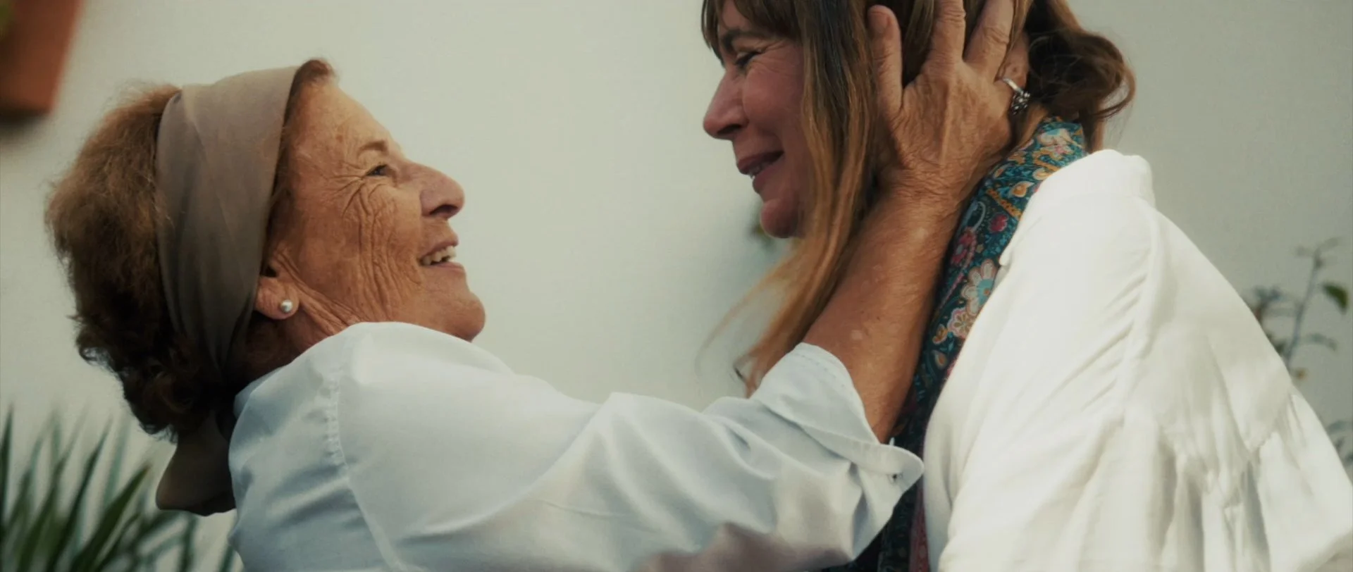 Two women, one elderly with a headscarf and the other middle-aged with long hair, smiling and embracing each other warmly.