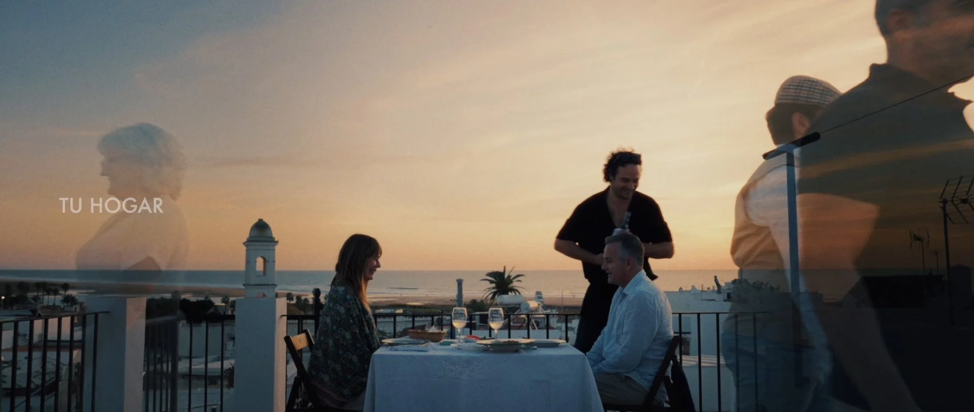 A couple dining at a table on a balcony during sunset, with a waiter serving them in Conil de la Frontera. The background features the ocean, a clear sky, and a sunset glow.