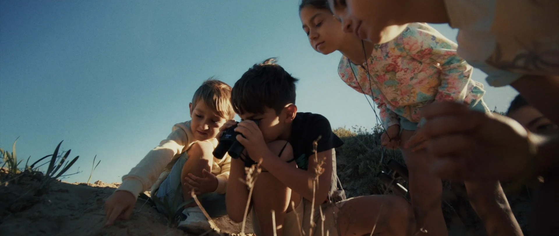 Four children, three girls and one boy, are outdoors in a natural setting, with the boy using binoculars to look at something on the ground. The other children are closely observing and leaning forward, with one girl pointing at the ground. They are 