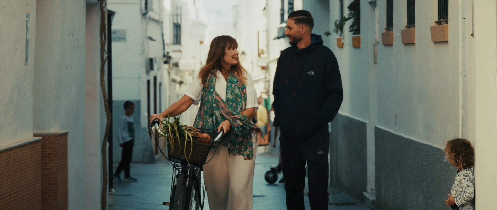 A woman with brown hair, wearing a white shirt and beige pants, is holding a bicycle and talking with a man with dark hair and a beard, wearing a black jacket. They are walking down a narrow street, smiling at each other. A young girl with curly hair