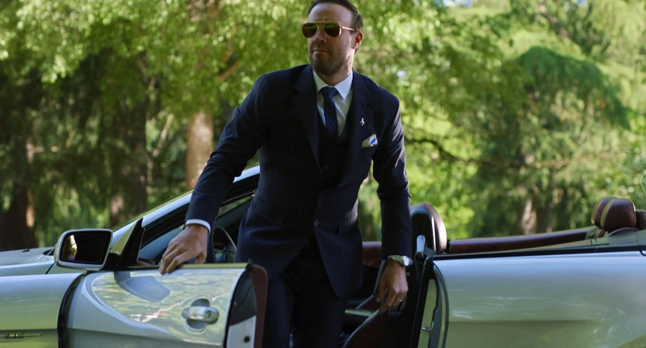 AB de Villiers con traje oscuro, gafas de sol, en un coche convertible, rodeado de árboles verdes en un parque o jardín.