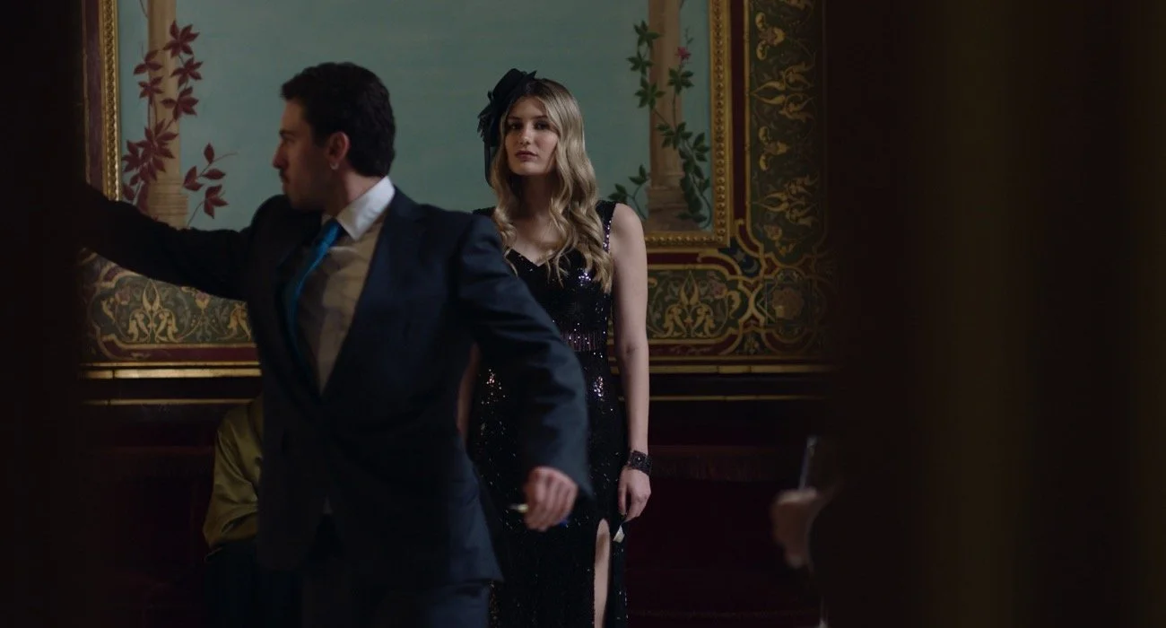 Una pareja en un evento elegante, la mujer lleva un vestido largo y negro con detalles brillantes y un sombrero pequeño, mientras que el hombre viste un saco oscuro con corbata azul, en un ambiente decorativo con arte en bares y columnas.