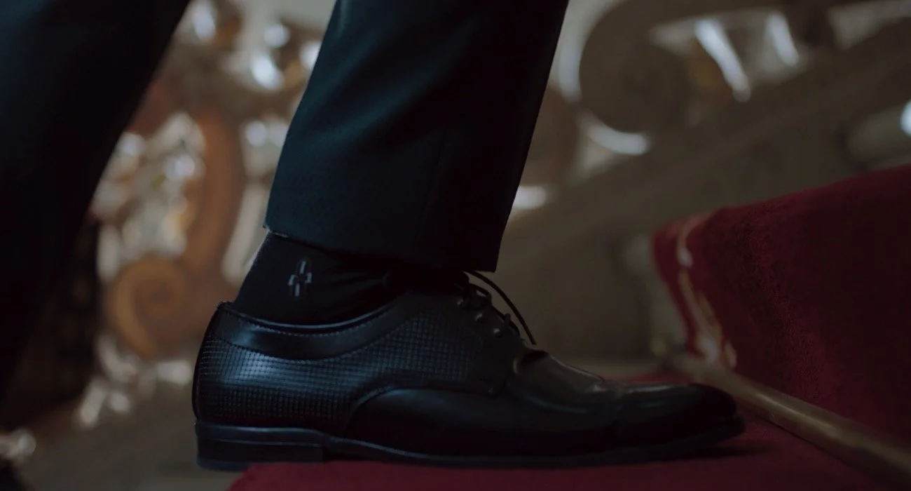 Zapatos formales negros con calcetas negras y pantalones oscuros, apoyados en una escalera de color crema con peldaños dorados y tapete rojo en el piso.