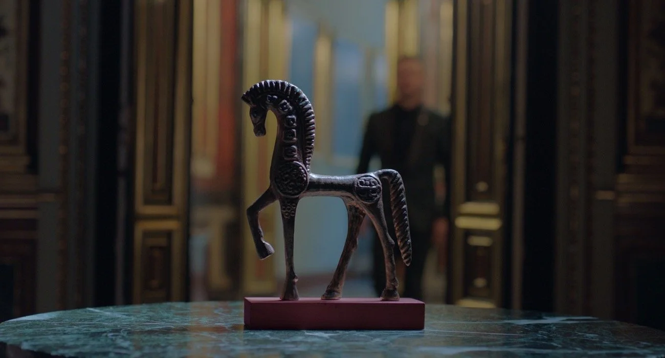 Una escultura de un caballo de color negro en una mesa, con un fondo de puertas decorativas y una persona en el fondo.