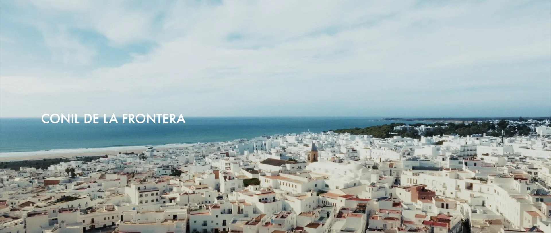 Aerial view of a coastal town with white buildings and a beach, labeled 'CONIL DE LA FRONTERA' on a cloudy sky.