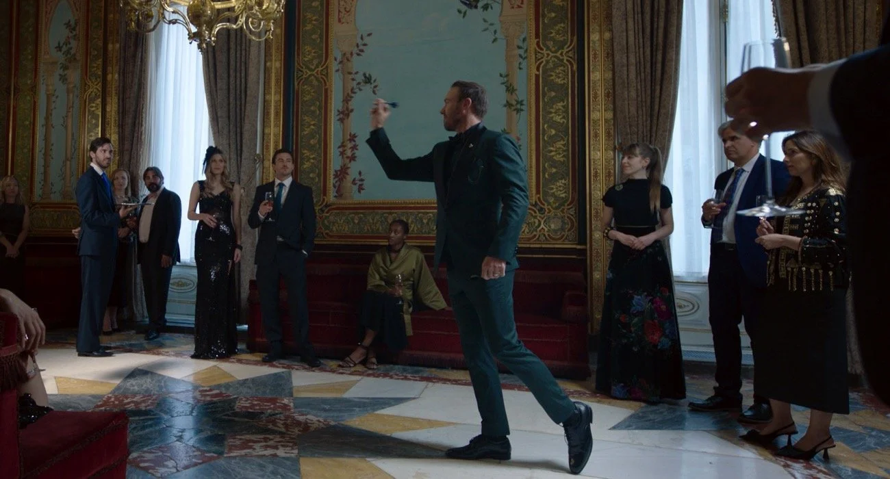 AB de Villiers en una fiesta elegante con varias personas observando y brindando en un salón decorado con cortinas y paredes ornamentadas.