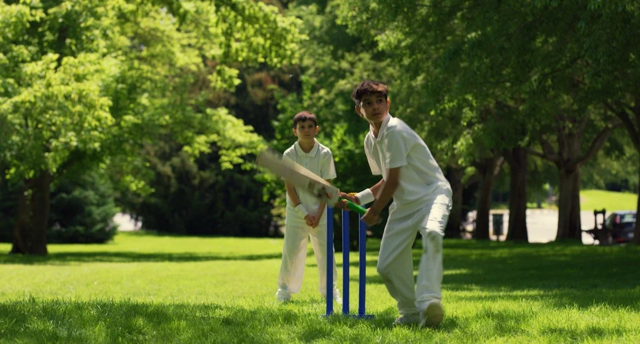 Dos niños jugando cricket en un parque con árboles verdes y césped