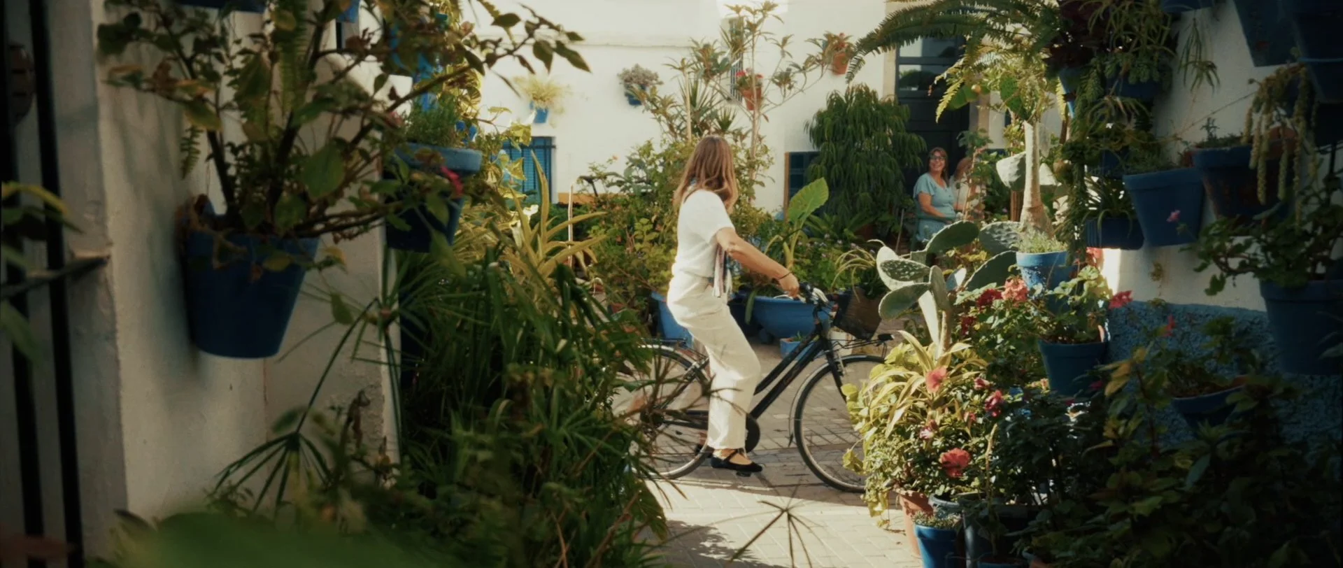 A woman riding a bicycle through a lush courtyard surrounded by potted plants and flowers, with another woman standing in the background near a white building.
