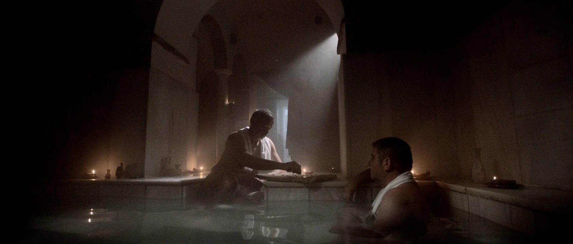 Dos hombres en una piscina o jacuzzi en un ambiente oscuro con iluminación suave y velas, uno está en el agua y el otro sentado en el borde, en un entorno que parece ser un spa o una sauna.