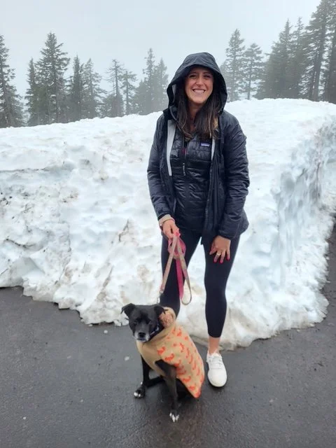 Woman in black rain jacket and leggings holding a dog in an orange coat, standing next to large snowbank on a wet road with evergreen trees in the background.