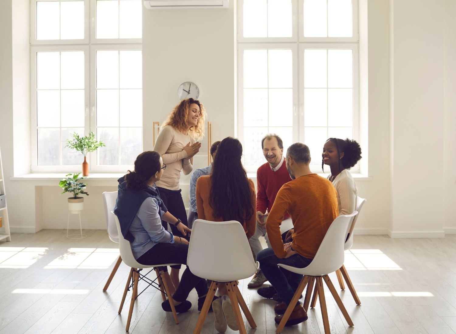 Group of diverse people sitting in a circle, smiling and engaging in conversation, in a bright room with large windows and sunlight.