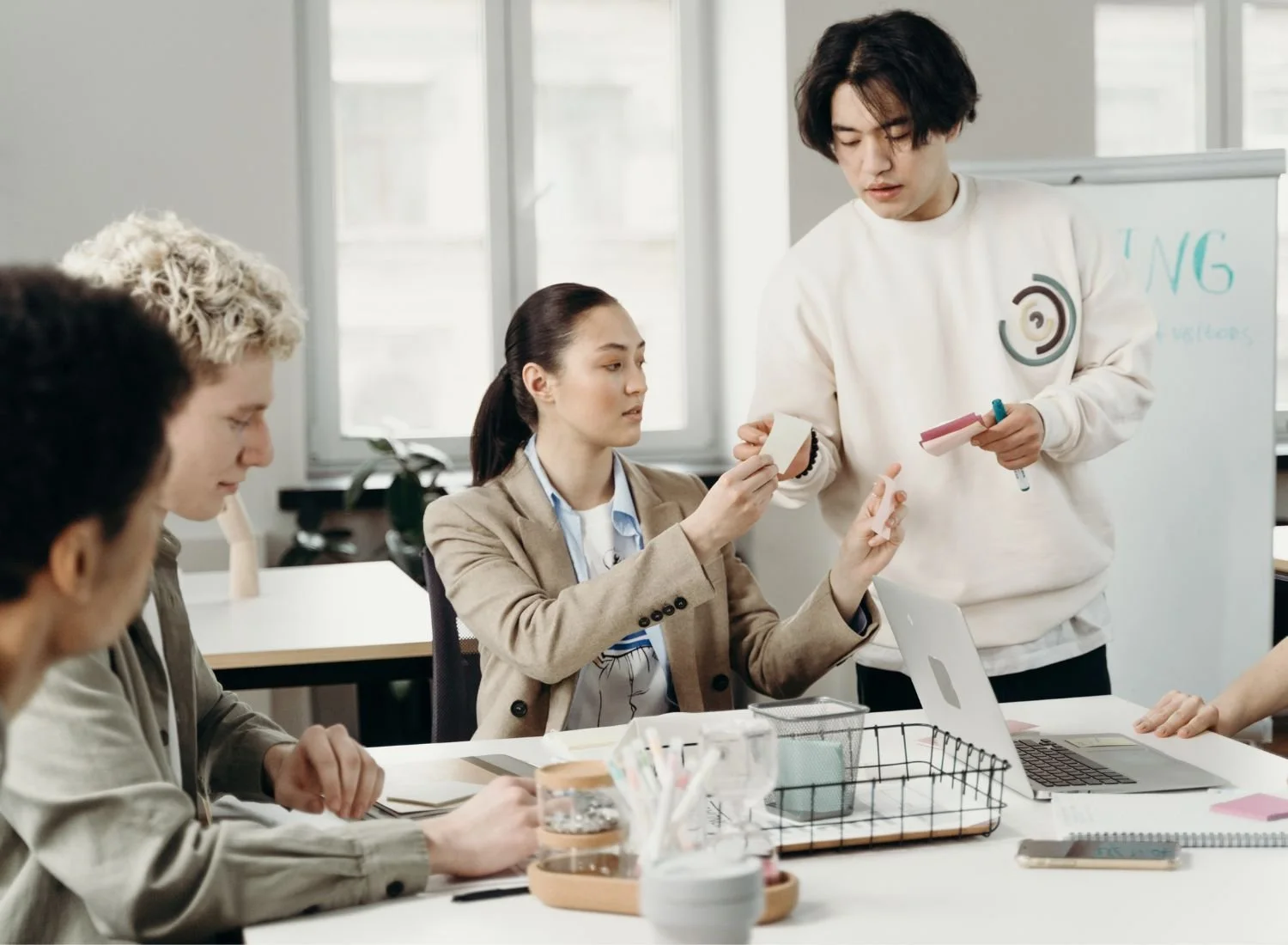 A group of five diverse young adults in a modern office or classroom, engaged in a presentation or discussion. One person is standing near a whiteboard, holding markers, while the others are seated, some taking notes and others listening attentively.