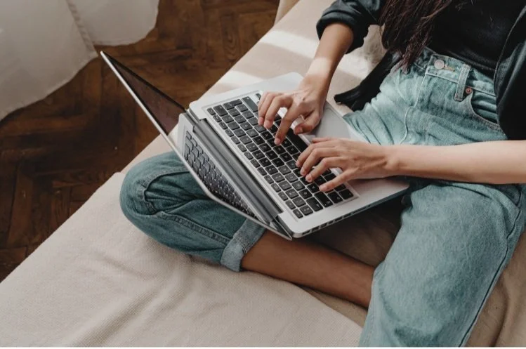 Person sitting on a couch using a laptop on their lap, wearing jeans and a black shirt.